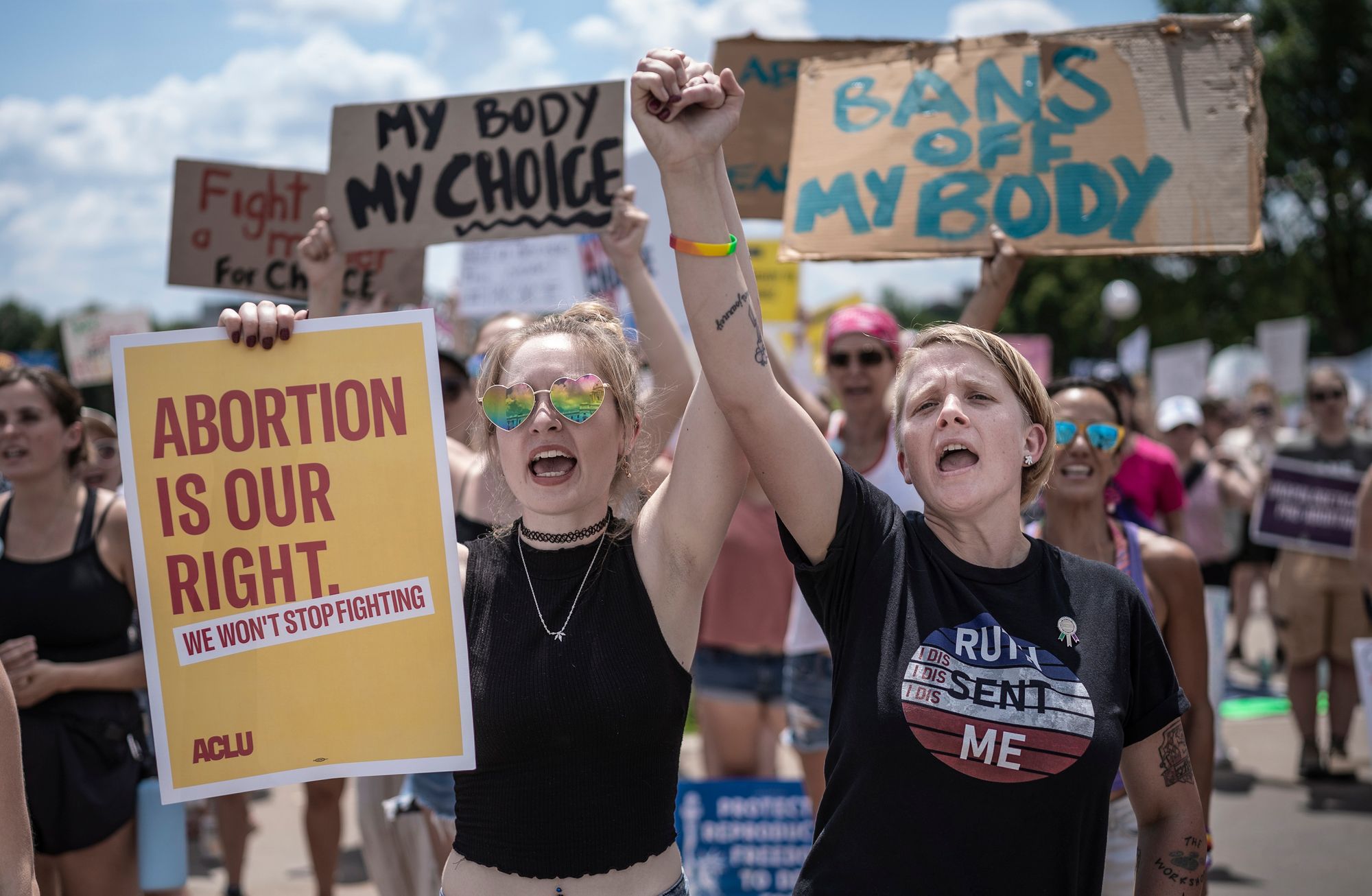 Two White people are shouting, standing in the foreground of a protest. A woman on the left holds a sign that says "Abortion is our right. We won't stop fighting." In the background, several individuals hold signs with phrases such as, "Bans off my body" and "My body, my choice."