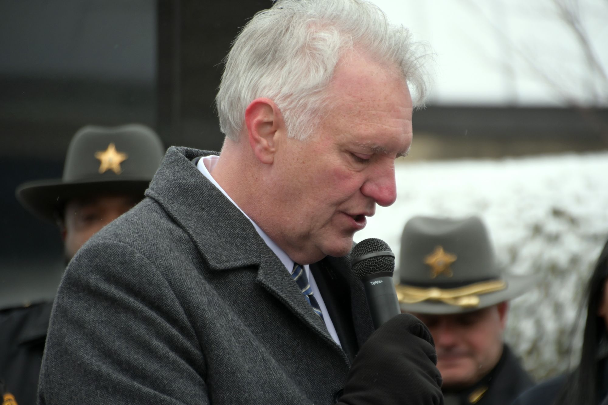 A White man with white hair speaks into a microphone outdoors, with two White men in uniform hats visible behind him.