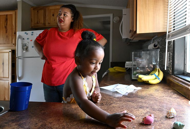 A young Black girl, wearing a yellow blouse, plays with seashells on a kitchen countertop. A Black woman, wearing a red blouse and red lipstick, stands with her hands on her hips in the background.