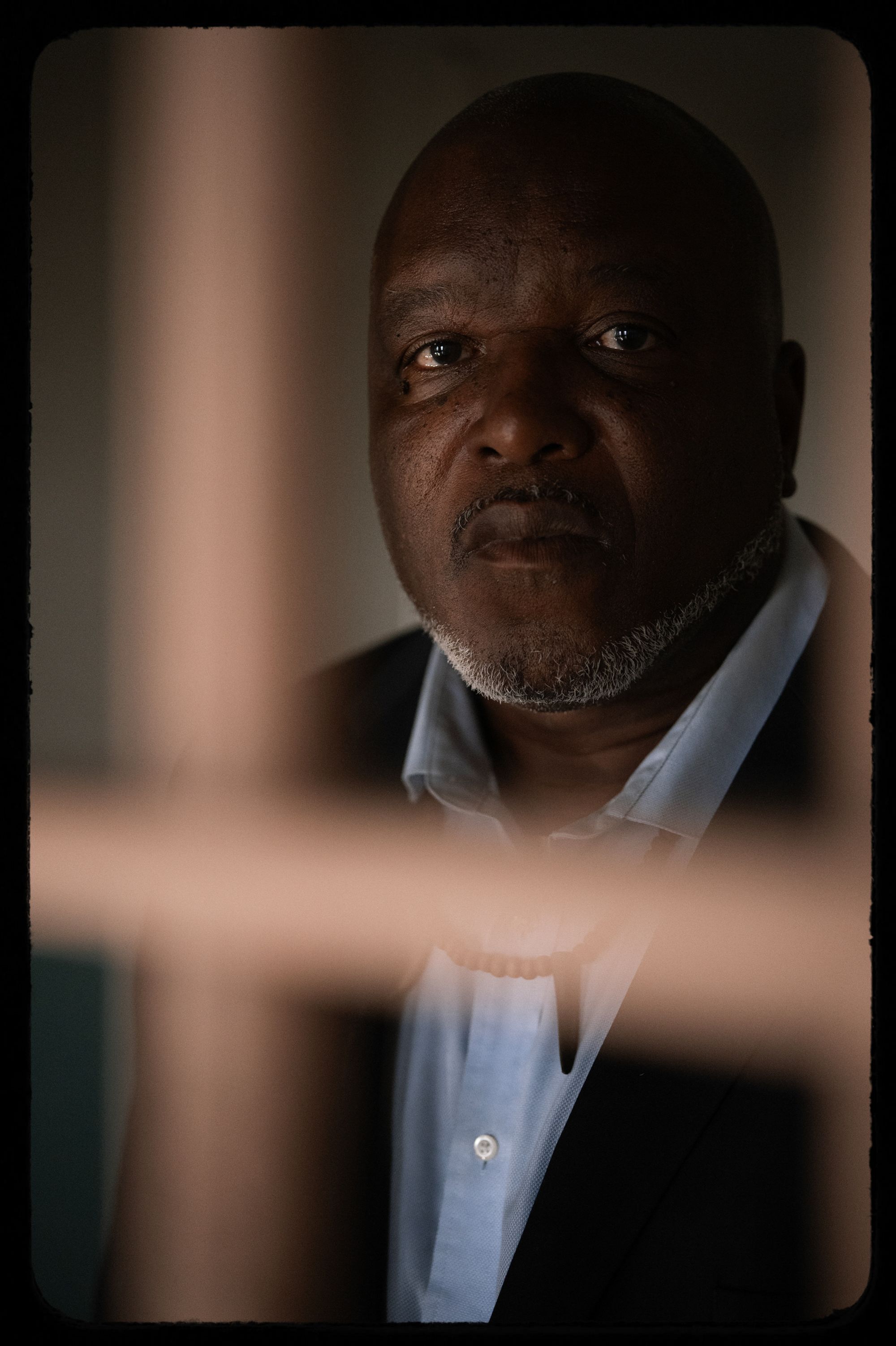 A photo shows Troy Williams, a Black man wearing a black suit and a light blue shirt, standing in an indoor space with bars in front of him at Alcatraz.