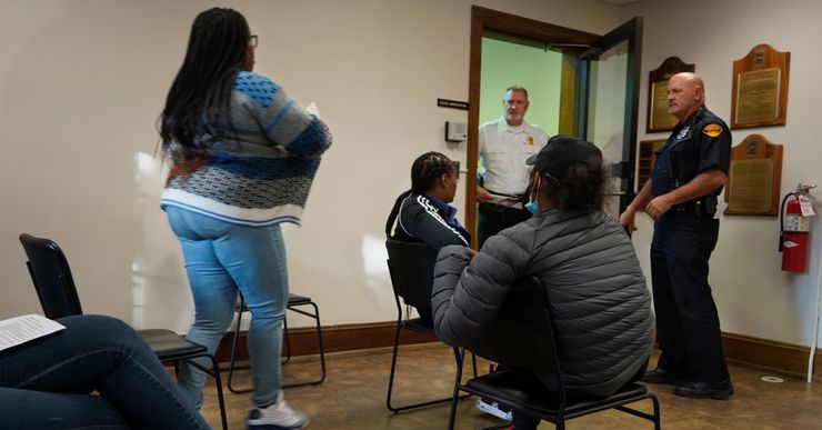 A Black woman stands to attend a hearing for a traffic citation in a waiting room.  A few other people sit nearby.  Two officers stand near the door.  