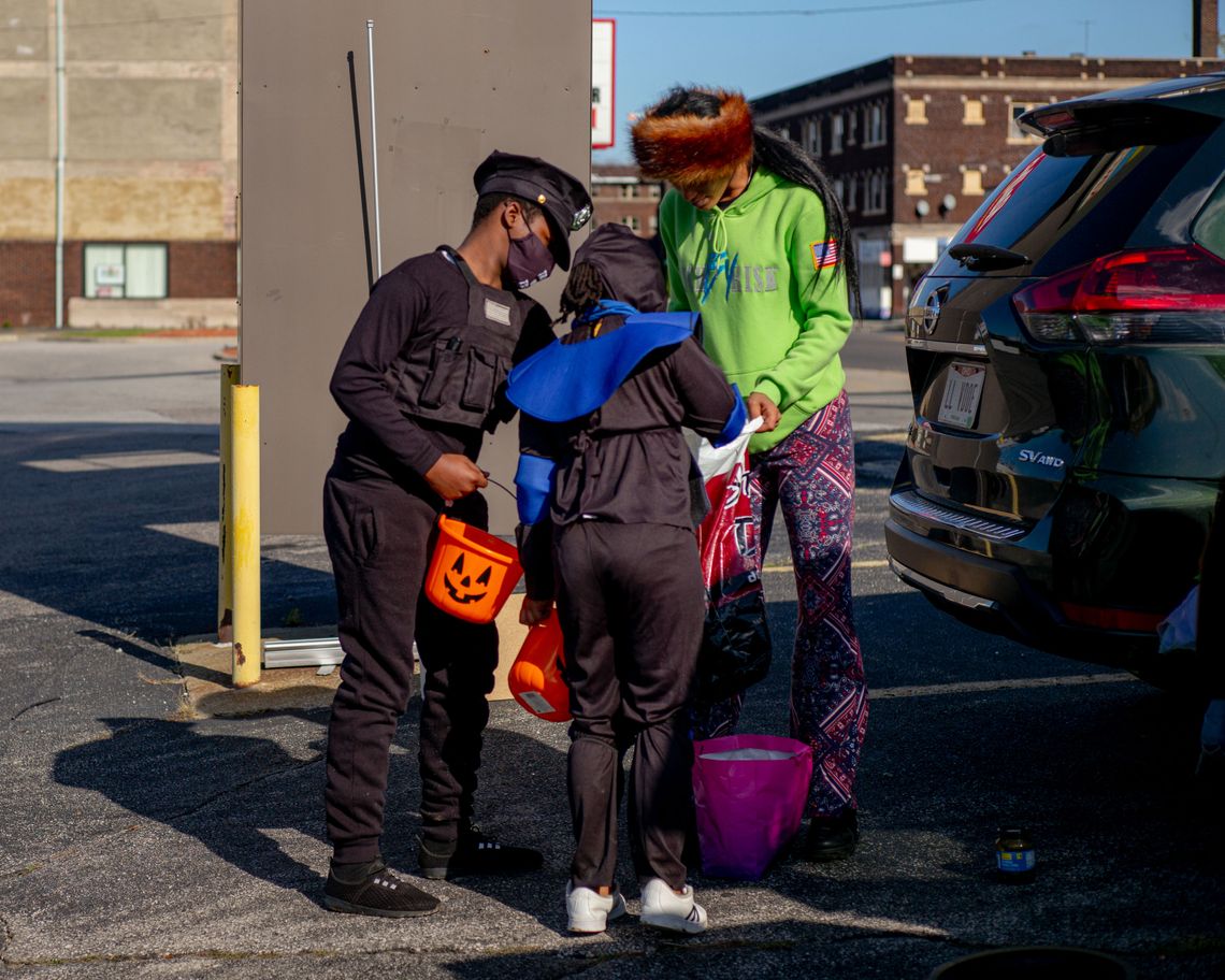 A photo of a Black woman wearing a bright green hoodie handing out candy to two children. One of the children is a Black boy dressed as a police officer while holding an orange Halloween bucket. 