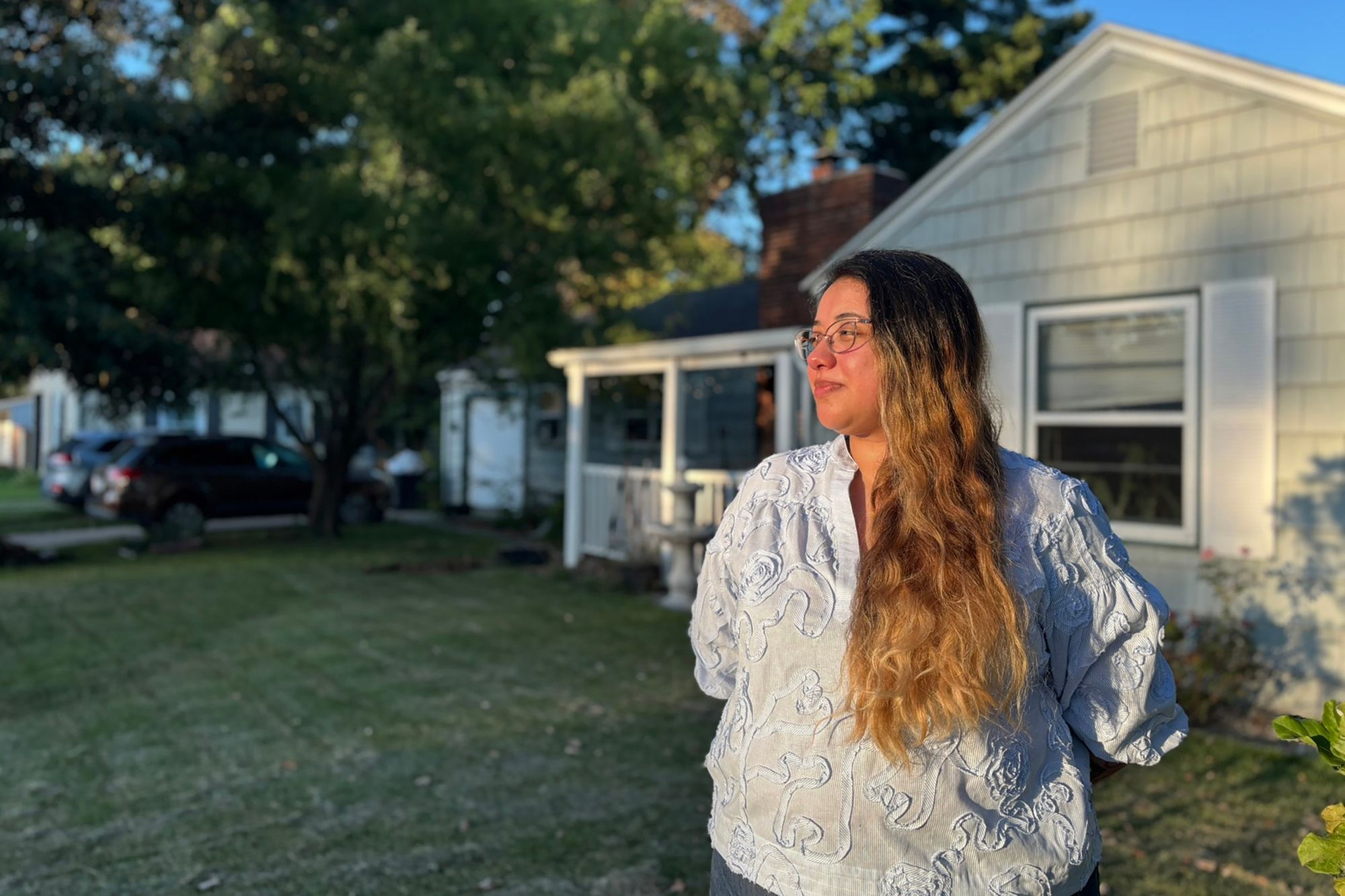 A light-skinned Latina woman with dark brown hair and blond highlights stands in a light blue blouse and blue jeans, with her hands behind her back, a small smile on her face, outside of a small light green house with a porch and a car sitting in the driveway.   