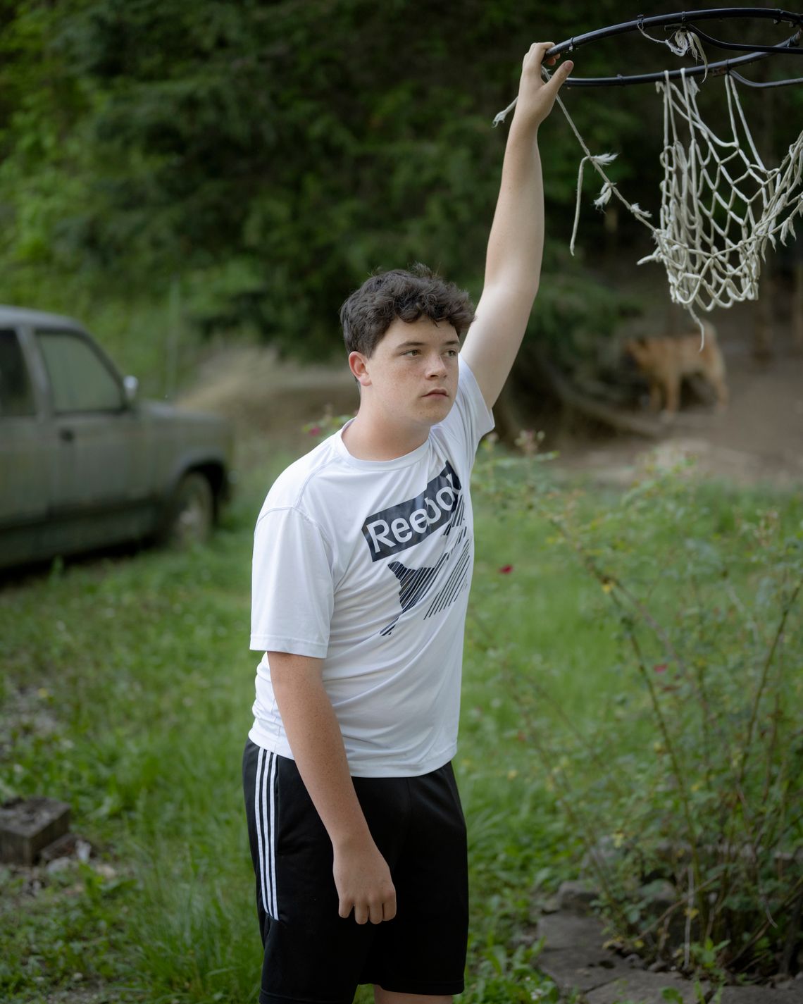 A White teenage boy wears a white T-shirt that says "Reebok" on it, with black basketball shorts. He is reaching up to hold onto a basketball hoop with a worn net and is outdoors surrounded by greenery, with a vehicle on the left.