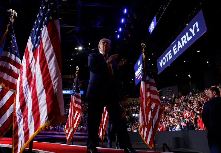 Former President Donald Trump, a White man in a red tie and navy suit, claps his hands as he walks off a stage during a rally inside an arena. He walks down a red rug between U.S. flags. In the background, there are signs that read "VOTE EARLY!" and hundreds of supporters sitting the stands.