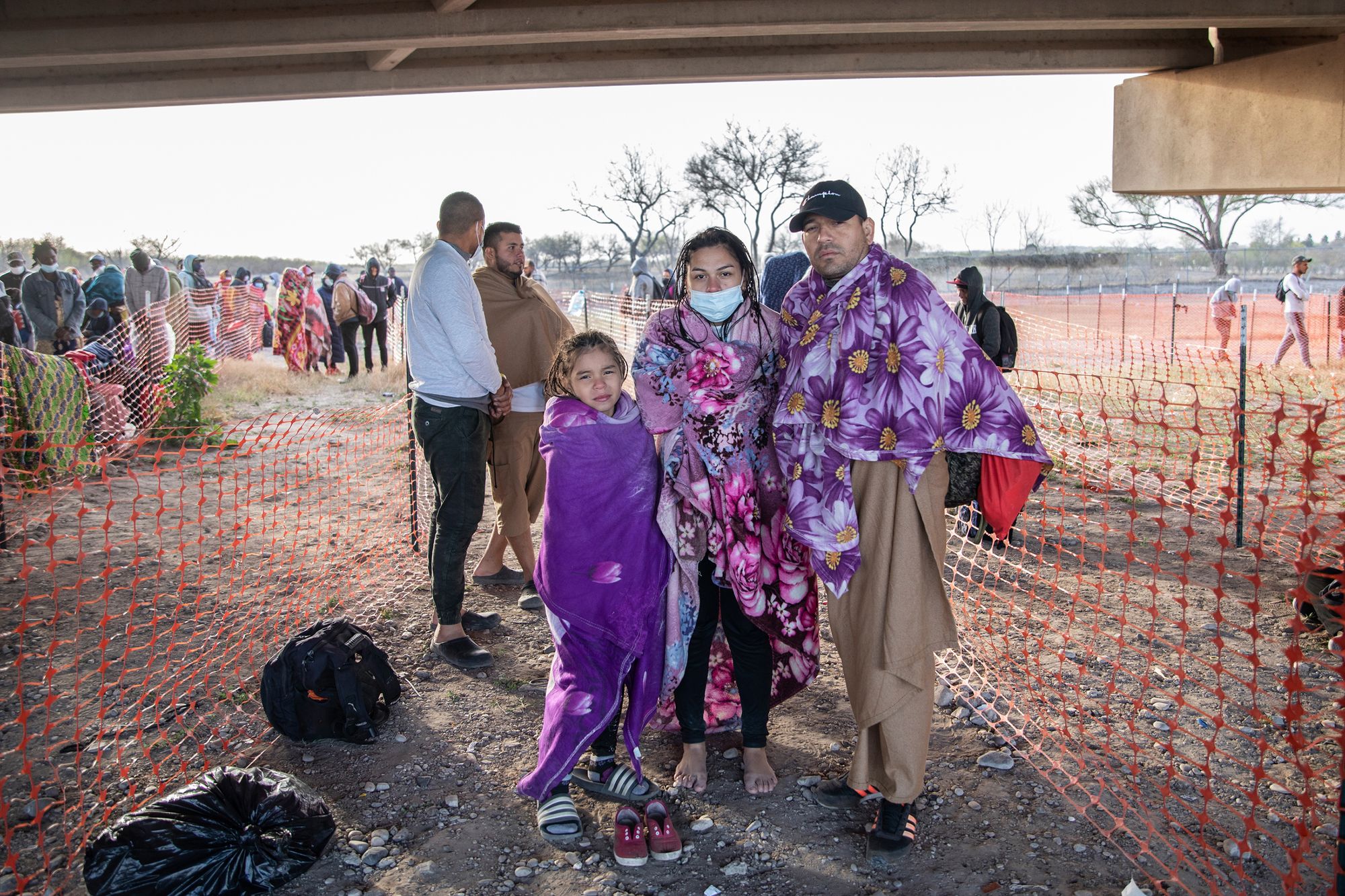 A young girl, her mother and her stepfather are facing the camera, wrapped in purple blankets. There is orange plastic mesh fencing for people to form lines behind them. 