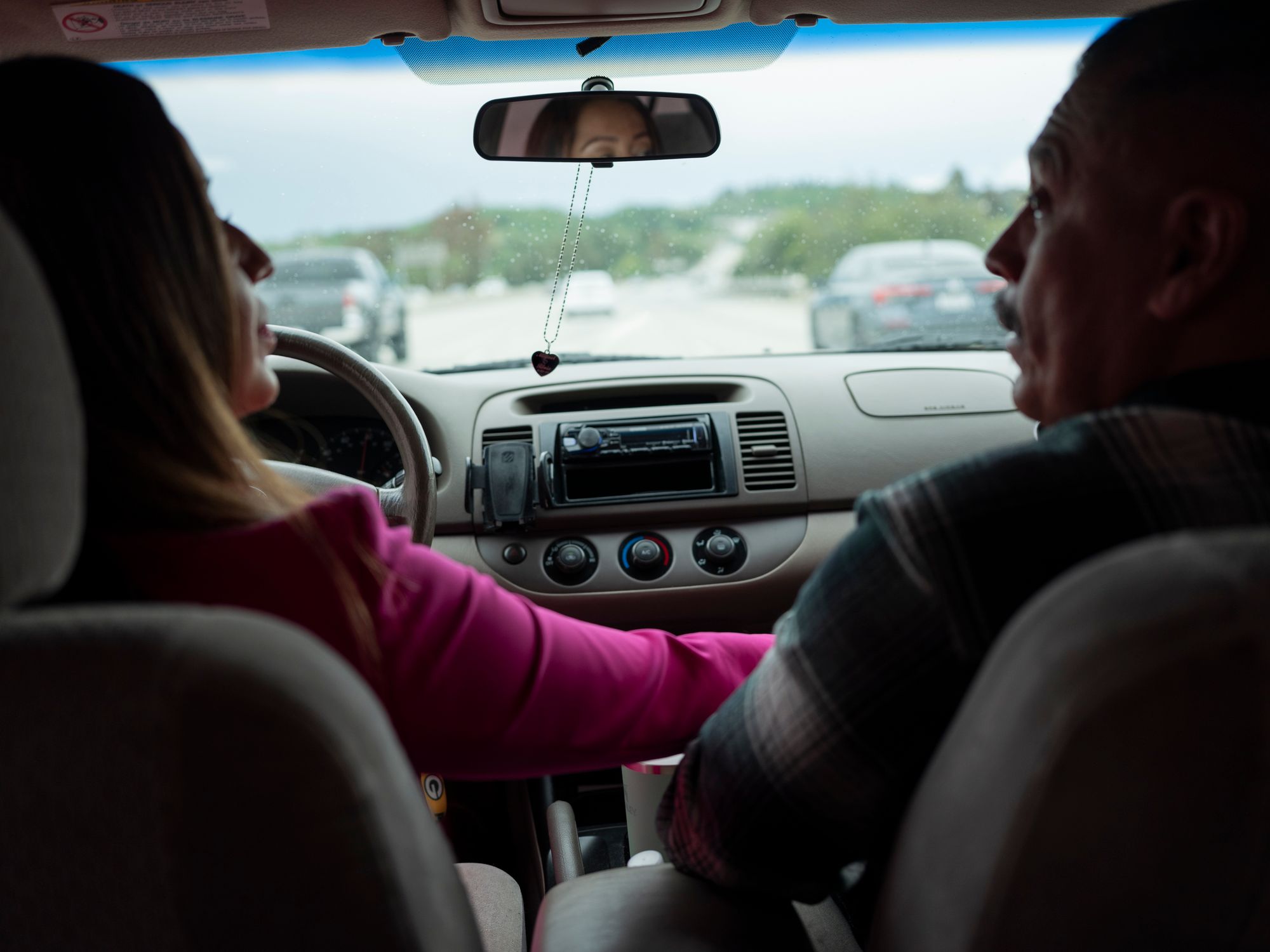 Silvia drives with her right hand holding Miguel's. A heart-shaped pendant hangs from the rearview mirror.