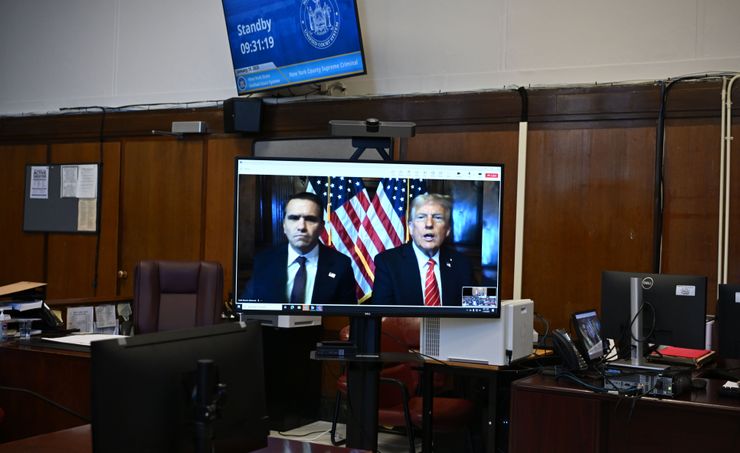 President Donald Trump, a White man wearing a dark suit and red striped tie, and his attorney Todd Blanche, a man with light-toned skin wearing a dark suit with a dark tie, appear on a monitor in a courtroom. 