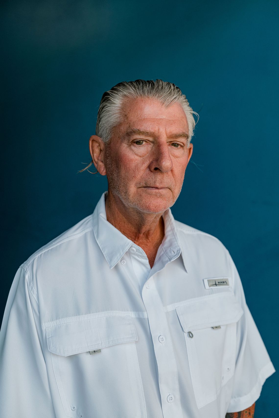 A photo of an older White man with slicked-back gray hair and a white button-down shirt stands in front of a blue background while looking into the camera.