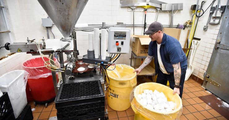 A man, wearing a blue prison uniform, white T-shirt, tan cap, and glasses, holds onto two yellow bins in front of an industrial ice cream machine. 