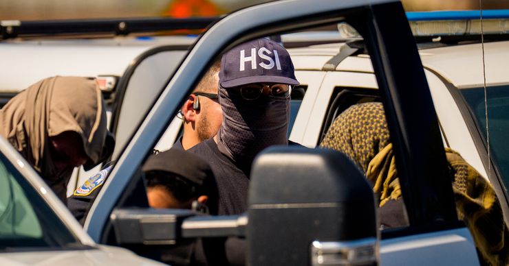 A person wearing a Homeland Security Investigations (HSI) cap has fabric wrapped around their face and is seen through the window of an open vehicle door at a checkpoint. To their left stands someone wearing a head covering, and to their right stands another person wearing a head covering. Someone else stands behind the person with the HSI cap.    