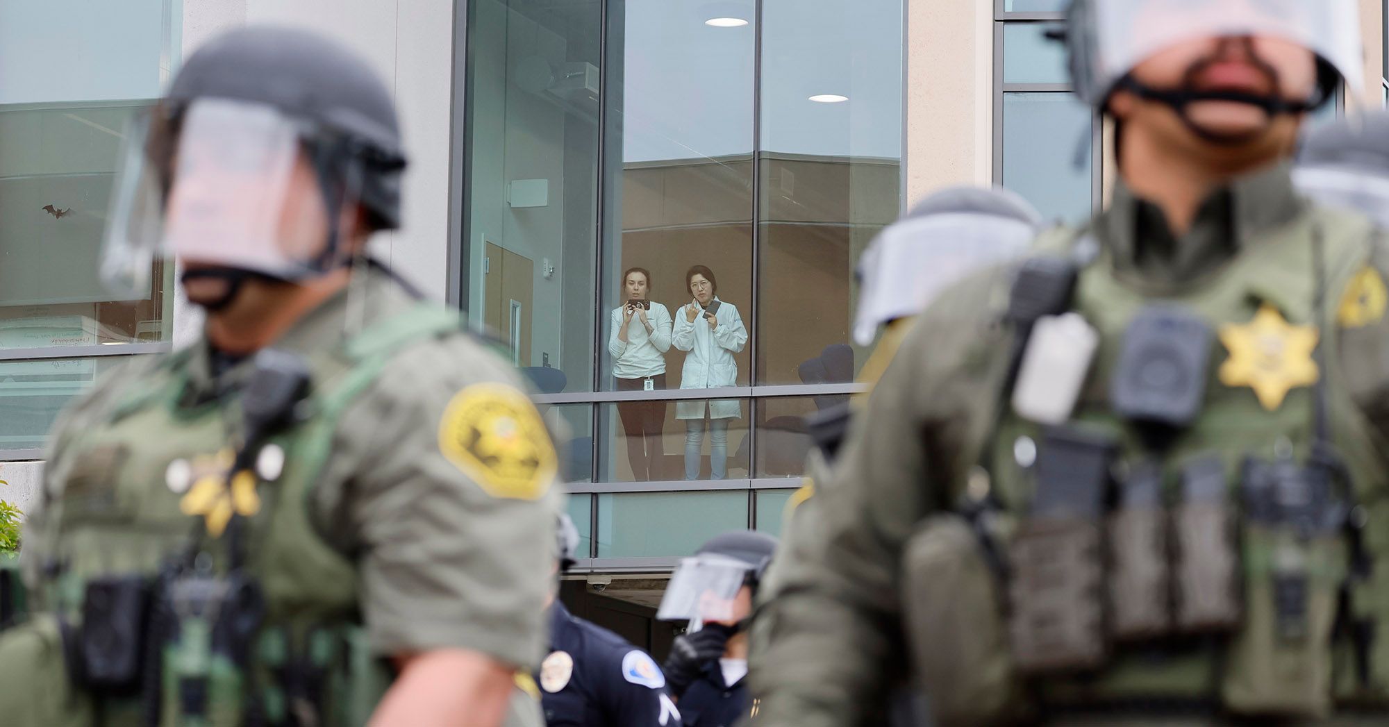 Two university members, wearing white tops, jeans and badges, look out a window and record videos from their phones. In the foreground, police wearing riot gear form a line in front of a building. 