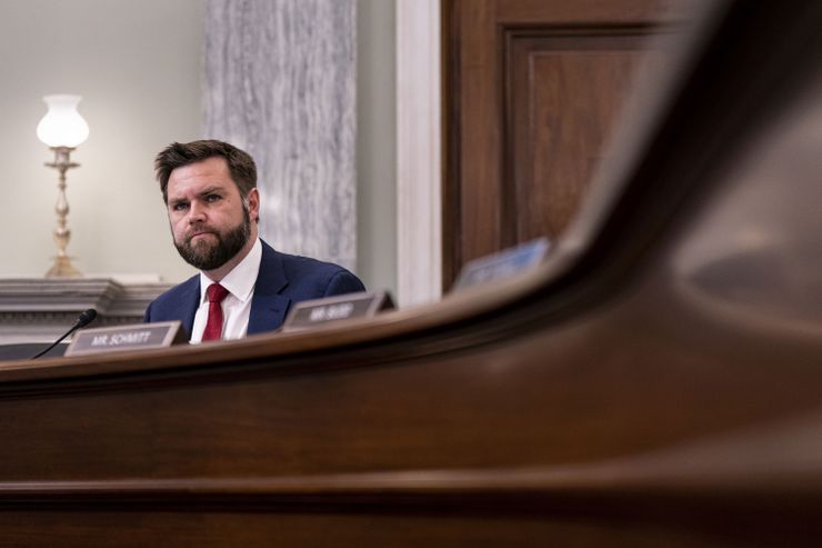 A middle-aged White man with brown hair and a beard, wearing a blue suit with a red tie, sits in front of a microphone at the end of a curved, wooden table. 