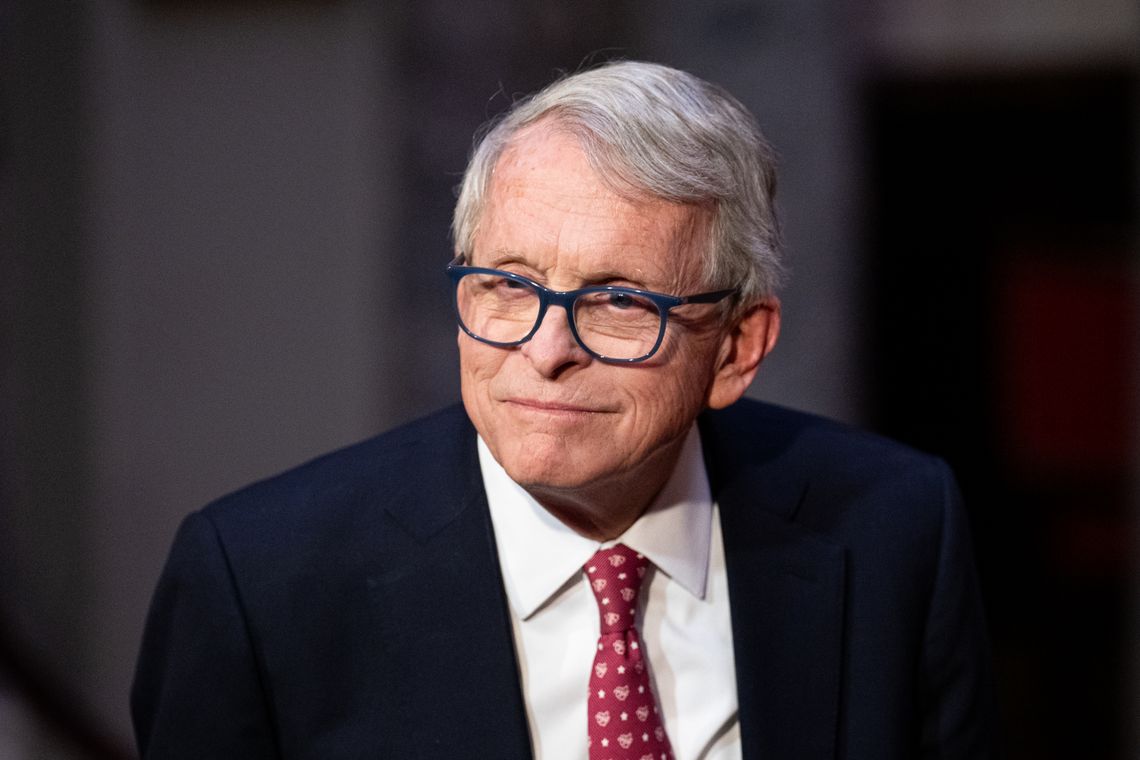 A photo shows a White man with white hair, wearing glasses and a dark suit jacket and red patterned tie. 