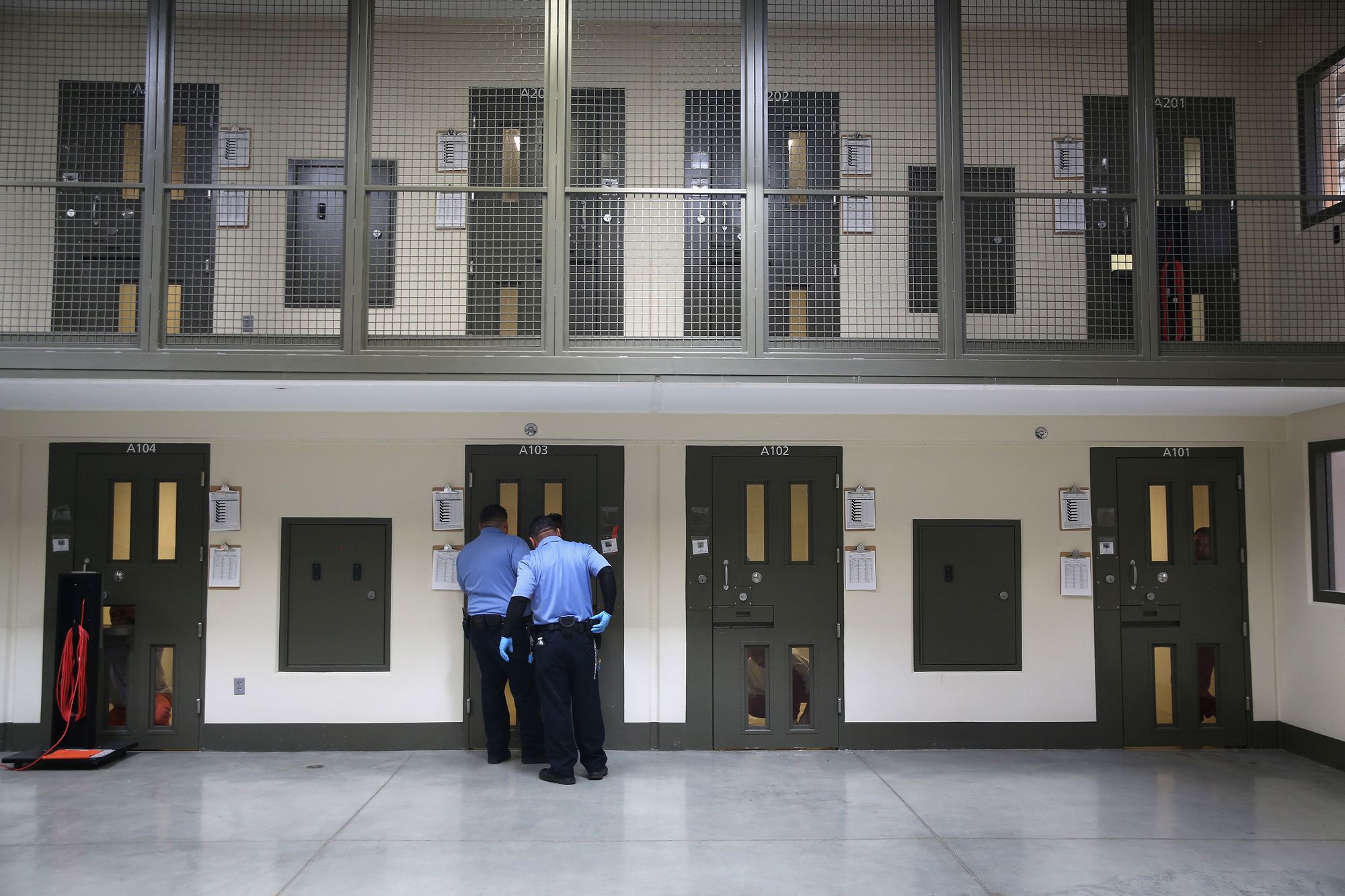 A photo shows the interior of an ICE Processing Center, where two floors are visible. There are four metal doors, each with four narrow vertical windows, on each floor. Two officers dressed in light blue shirts and dark pants stand at one of the doors on the first floor.  
