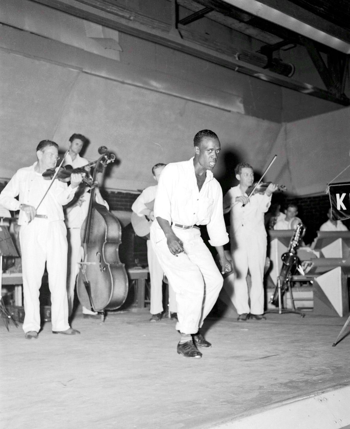 A man faces the camera while dancing in front of a group of musicians playing instruments. 