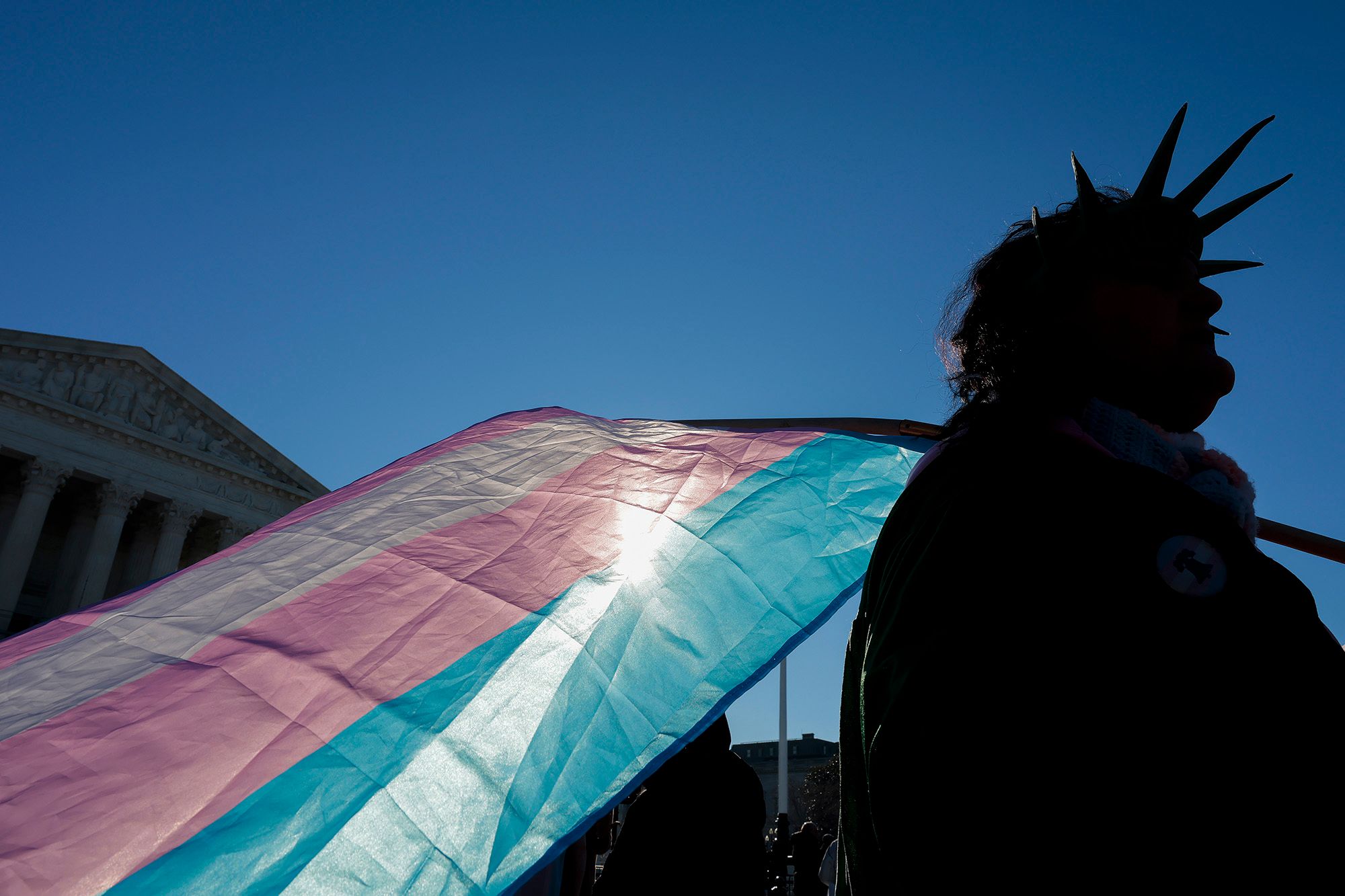 A photo shows a silhouette of a person, wearing a Statue of Liberty crown, holding a transgender pride flag. The U.S. Supreme Court building is in the background. 