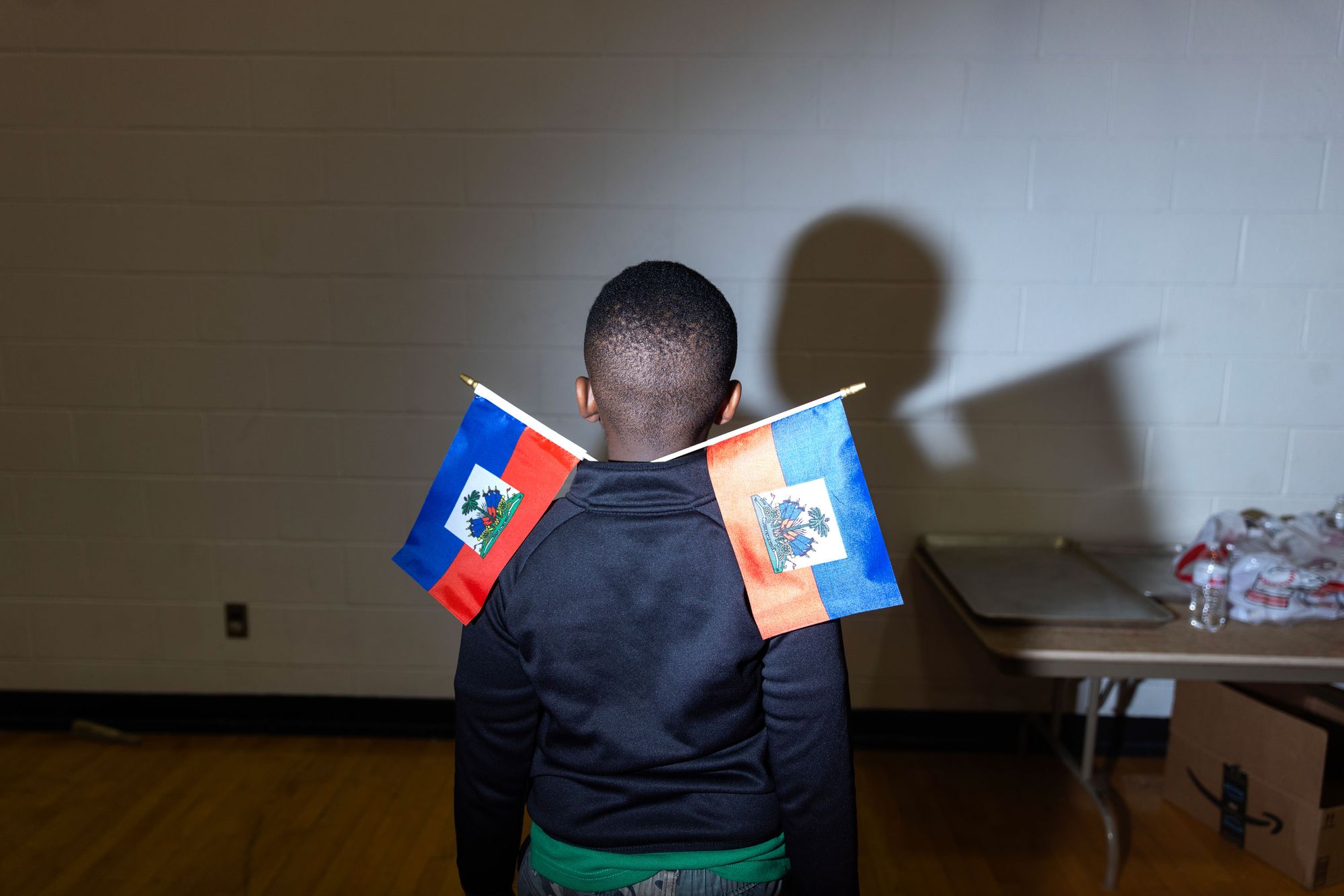 A photo shows a Black boy turned away from the camera with two Haitian flags sticking out of his shirt collar. 