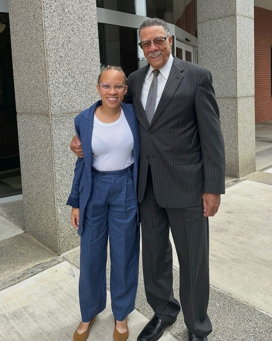 A photo shows a smiling Black woman in a blue pantsuit next to a smiling Black man in his 80s wearing a dark gray suit. 