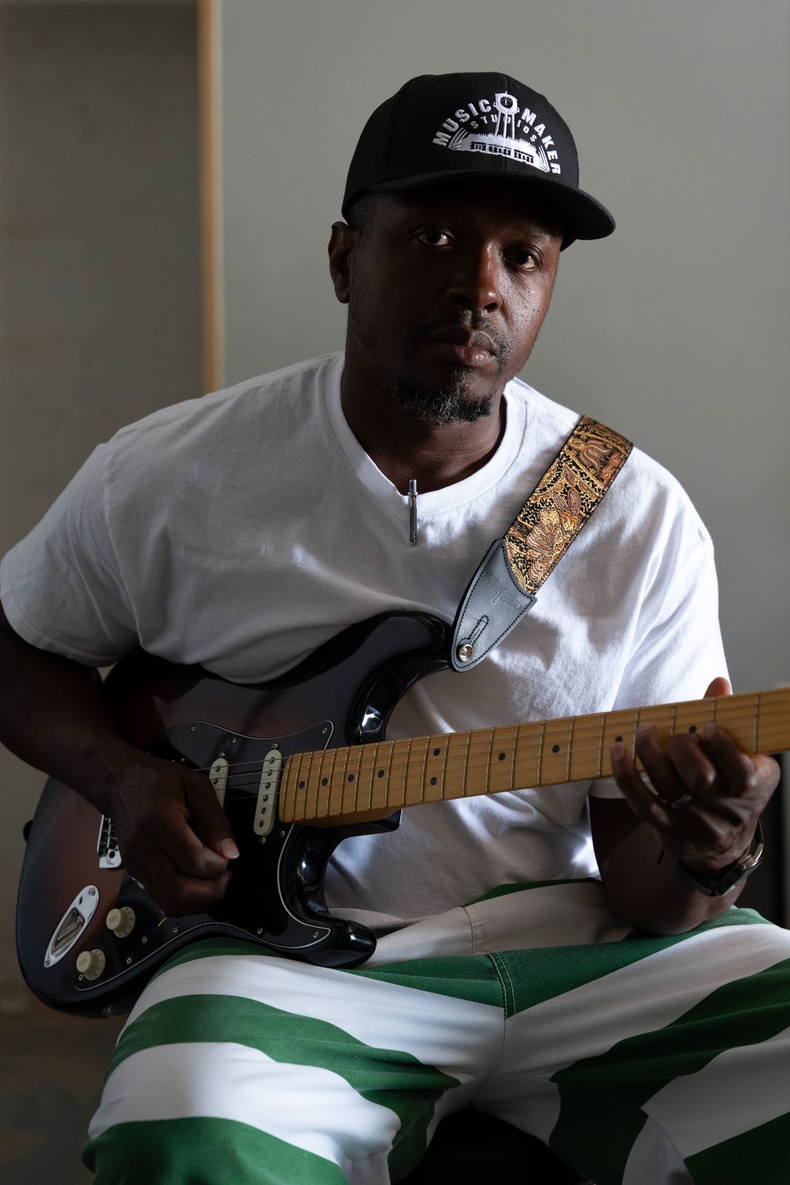 A photo shows a Black man wearing a white T-shirt, green-and-white striped pants, and a black baseball cap with the words “Music Maker Studios” on it. He is holding an electric guitar.