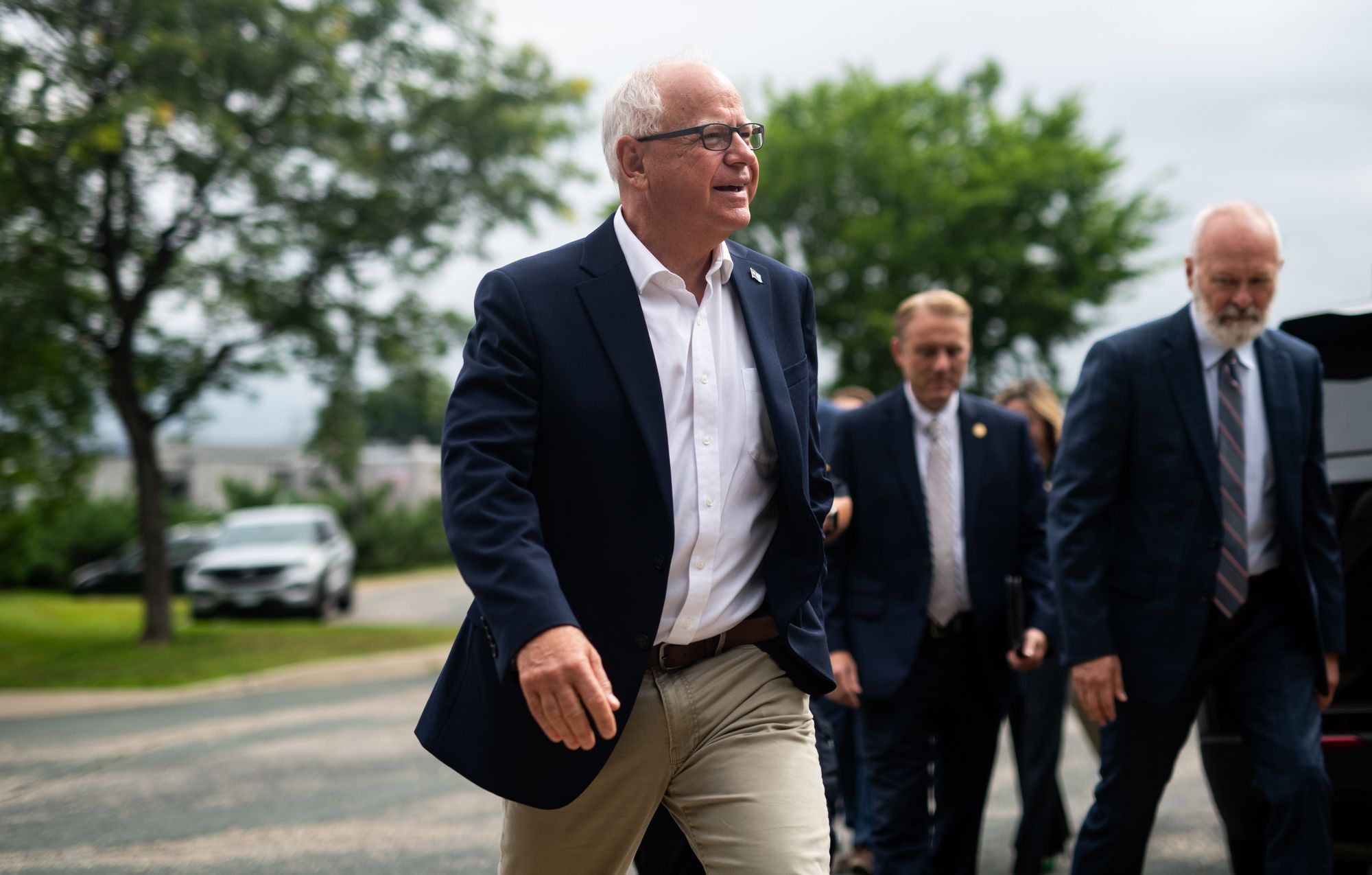 Tim Walz, a White man with white hair and glasses, wearing a navy suit jacket and khaki pants, walks outside with two men behind him to his left.  