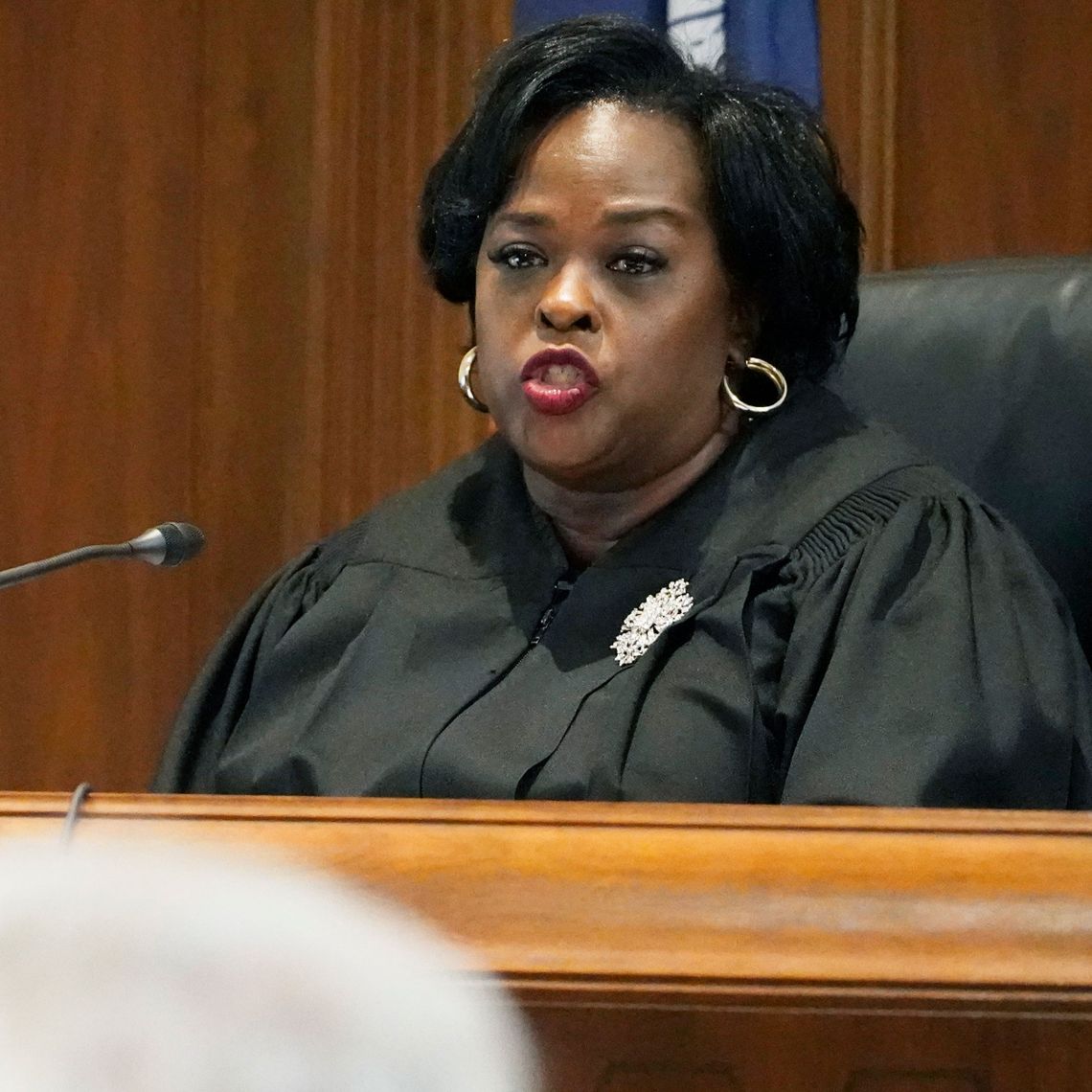 A Black woman in a black judge's robe speaks on the bench in a courtroom. 