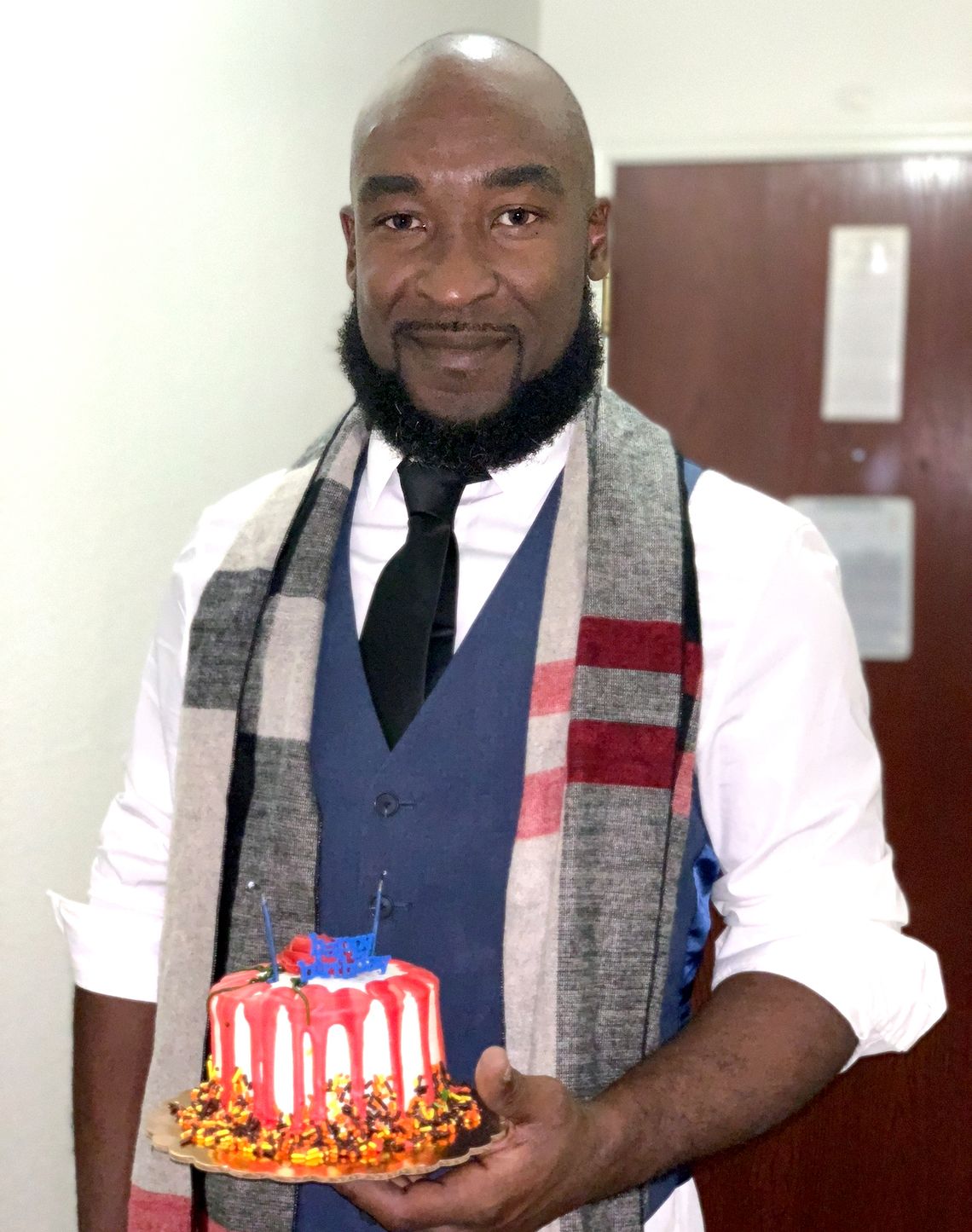 Rodney Taylor, a Black man, wears a white long-sleeved shirt, a blue vest, and a gray scarf around his neck, while holding a small cake that is red, white and blue in color. 