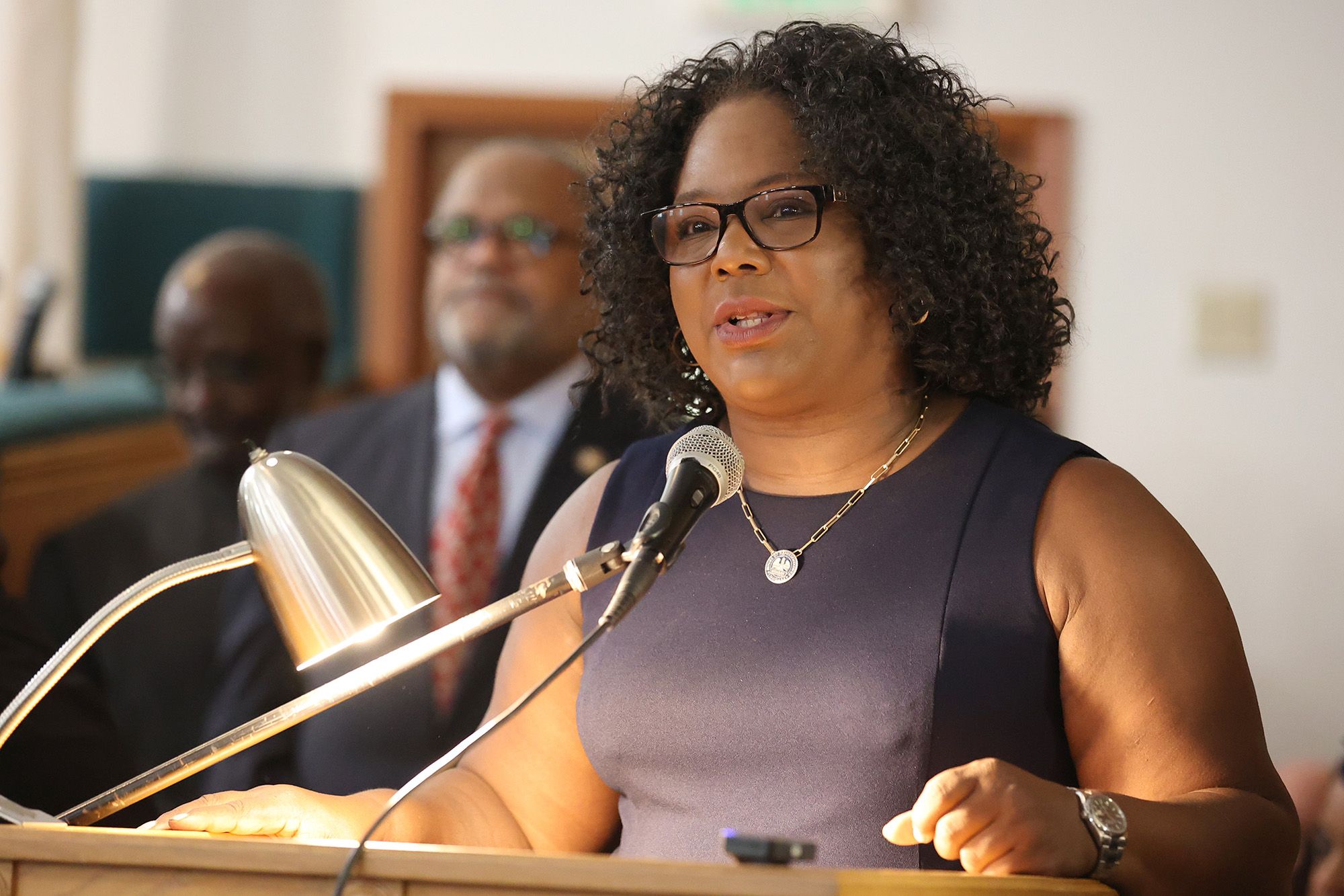 Louisiana Rep. Delisha Boyd, a Black woman wearing glasses and a black dress, speaks into a mic while standing in front of a podium. Two Black men wearing suits stand in the background. 