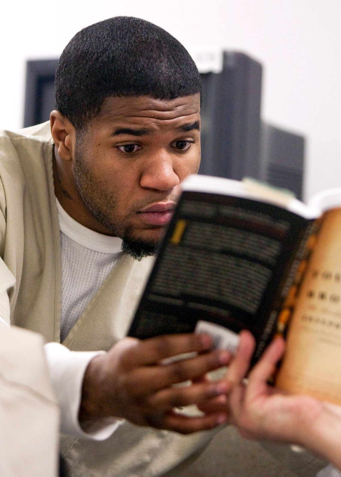 A Black man in a tan prison uniform reads a book, which is being held by his hand and a medium-light toned hand. 