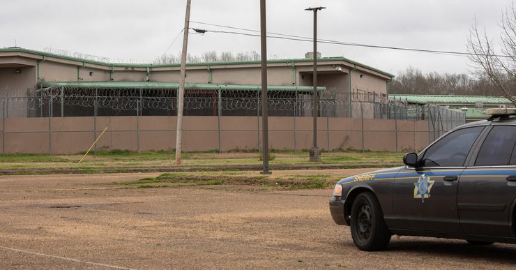 A vehicle with the word "Sheriff" on it is on the right side of a photo, while a facility surrounded by a barbed wire fence is on the left. 