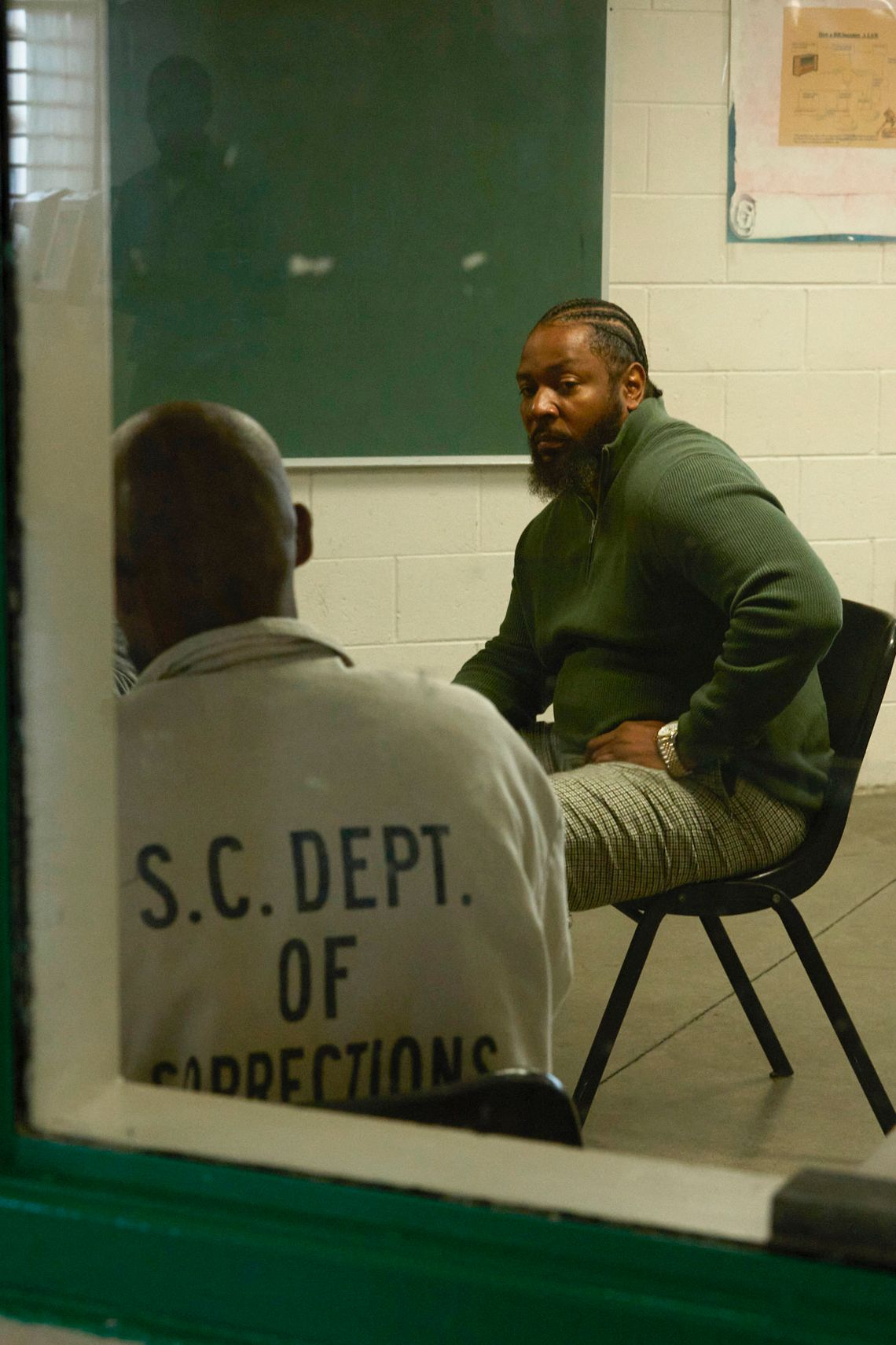 Through a window, Ofonzo Staton, a Black man with cornrows and a beard and goatee, wearing a dark green zip-up sweater and checkered pants, is visible. He sits in a chair next to a man wearing a shirt that reads “S.C. Dept. of Corrections” on the back.