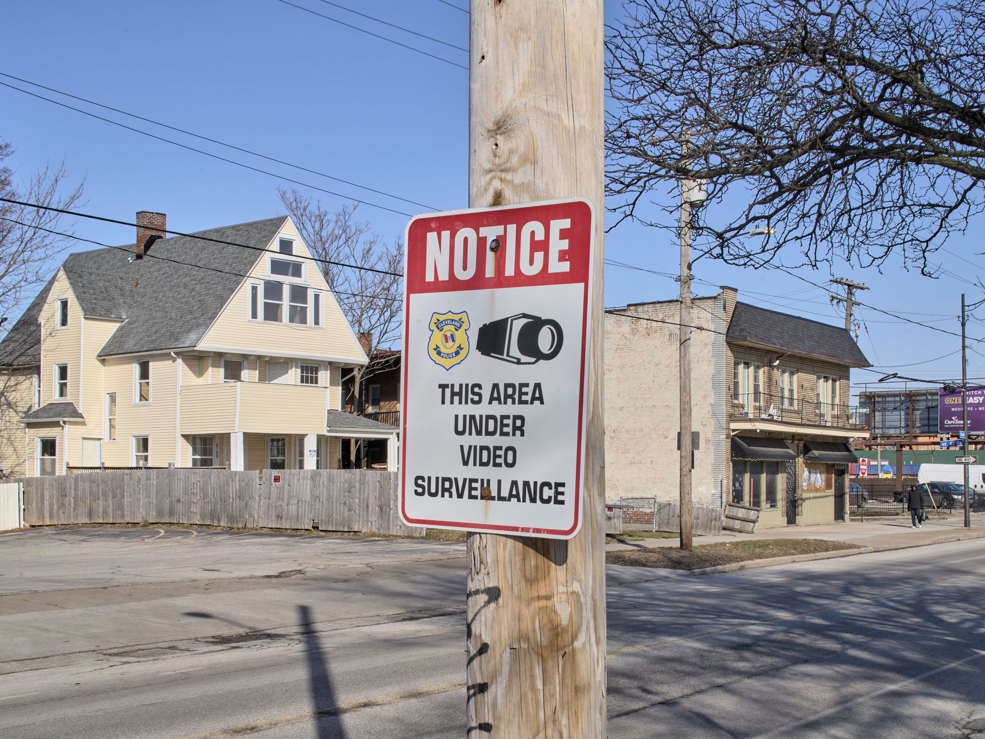 A photograph of a sign mounted on a pole in a residential area. The sign shows an illustration of a video camera and the Cleveland Police logo, and reads “Notice: This area under video surveillance.” 