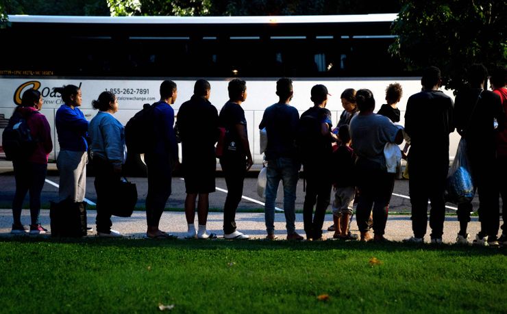  A group of about 15 people stand in line with their backs facing the camera, in front of a bus.