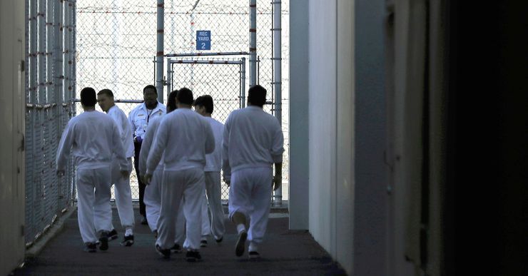 A group of men wearing white uniforms, seen from the back, walk toward a fenced area. A guard stands by the entrance. 