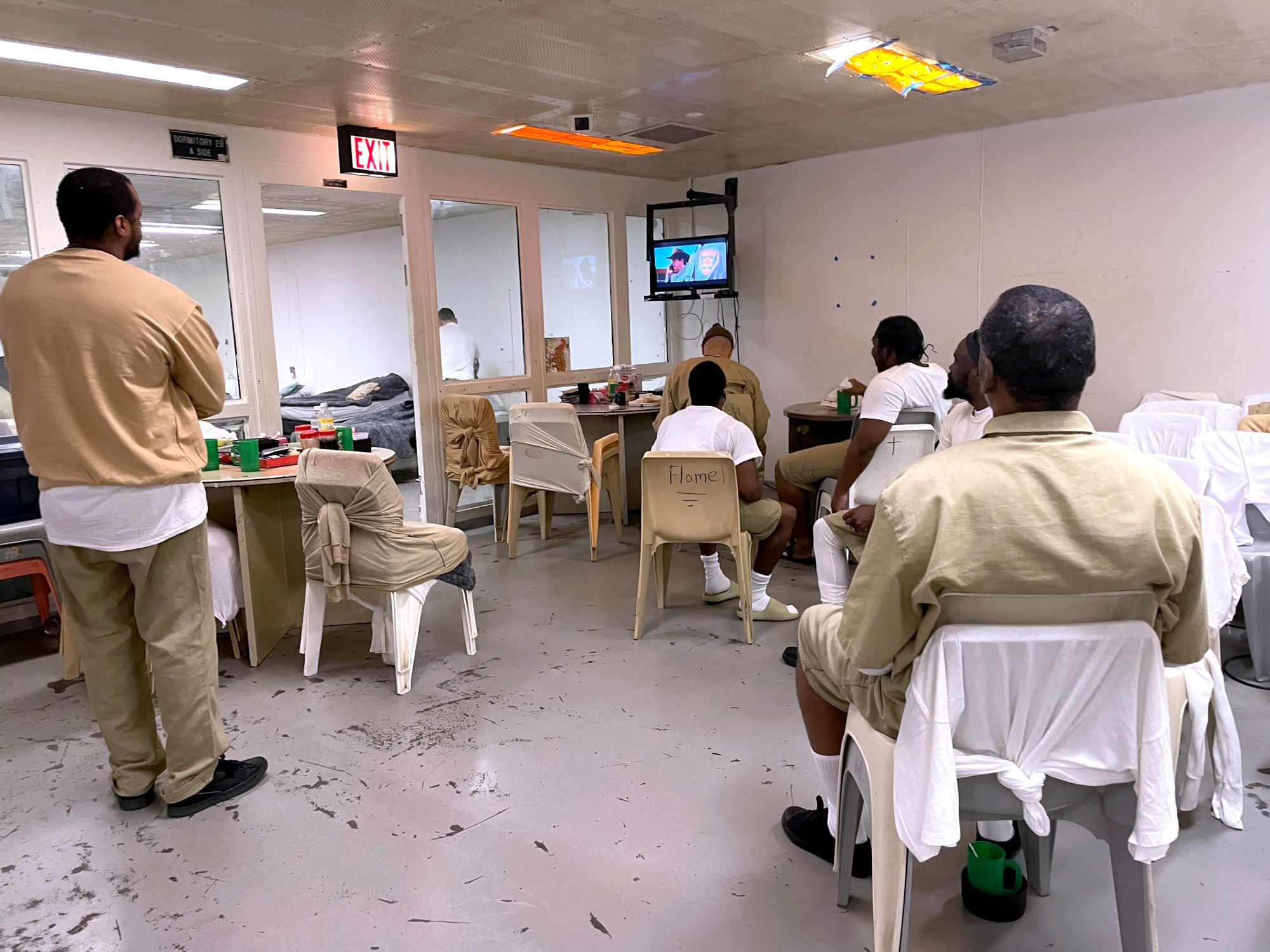 Men in tan prison uniforms watching TV. Wet uniforms are tied to the chairs to air dry. 