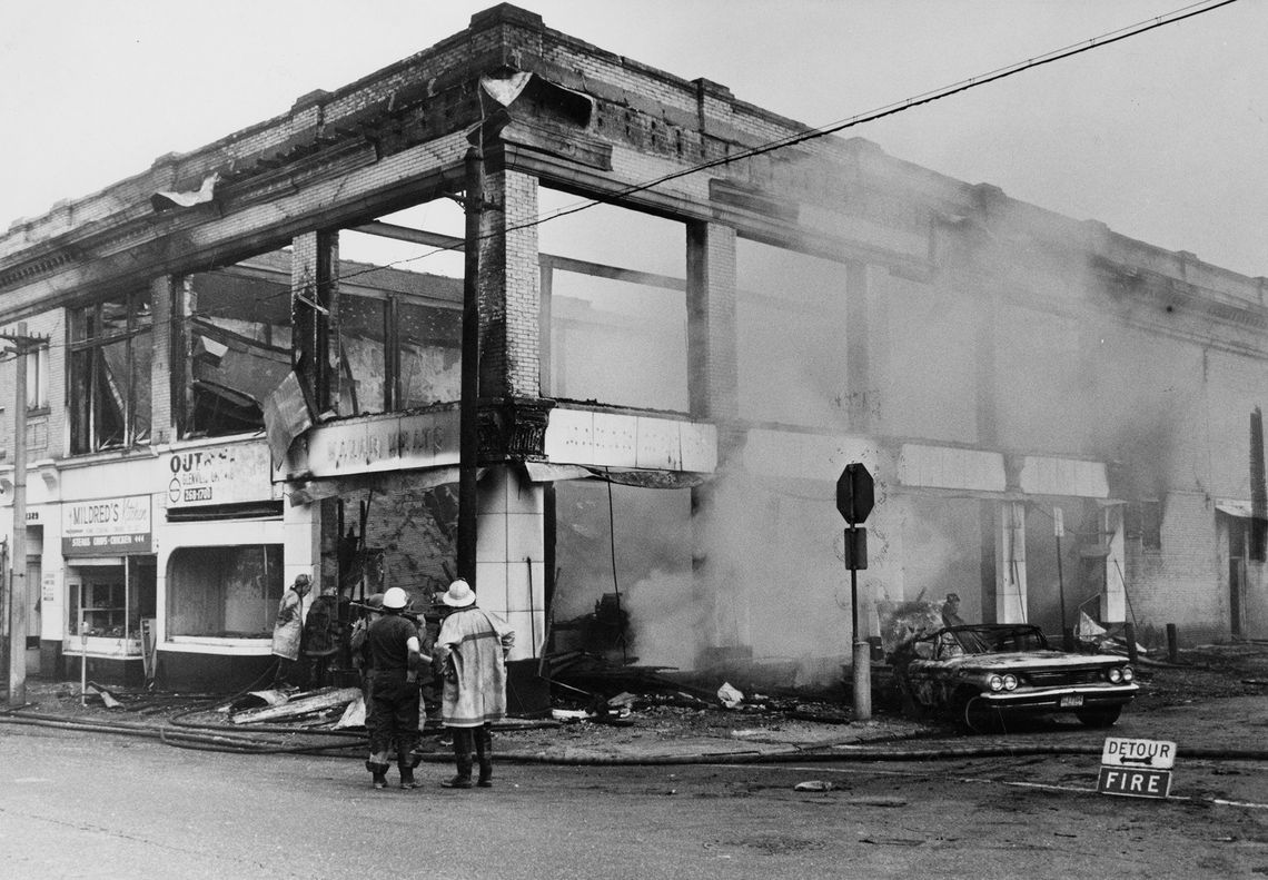 Two firefighters stand in front of a partially collapsed, smoking building, spraying water onto the flames. The scene is chaotic, with debris scattered on the ground and a damaged car nearby.