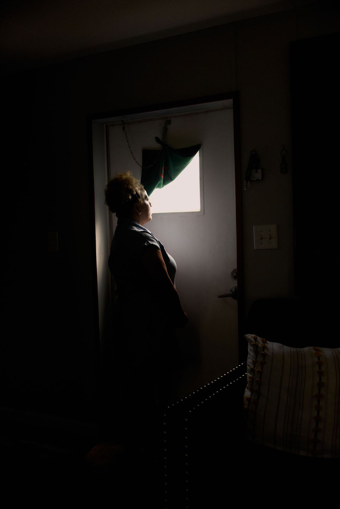 A photo shows a medium-skinned woman with blond curly hair in an updo, standing in her home while looking out a small window in a door. The inside of the home is dark. 