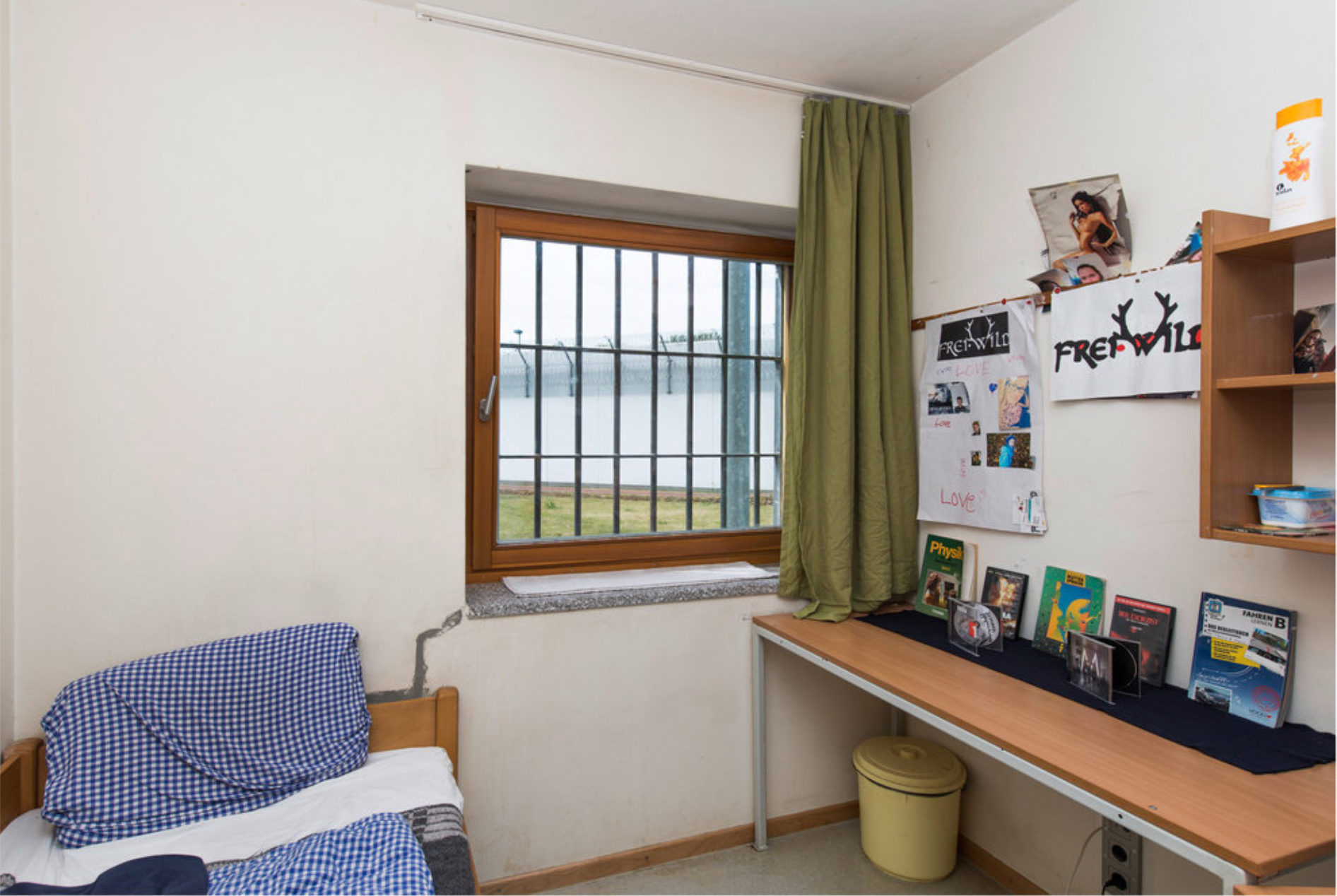 A room with white walls has a bed in the lower left corner with a pillow covered by a blue-checkered pillow case. On the right, is a long narrow table, which has a few books and CDs on it. Above that are two posters, both of which say “Frei Wild”, and one of them has antlers coming out of the two sides of the “W” in “Wild”. There are vertical bars on the window. 