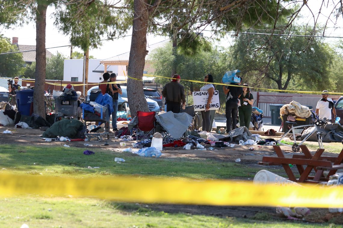 People stand around the remains of an encampment of unhoused people. People's belongings, a shopping cart, debris and camp chairs are visible on the site. 