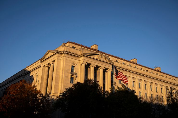 A color photo shows a gray stone building with columns. Dusk lighting makes the building appear yellow-orange. In the foreground are trees and an American flag on a pole.