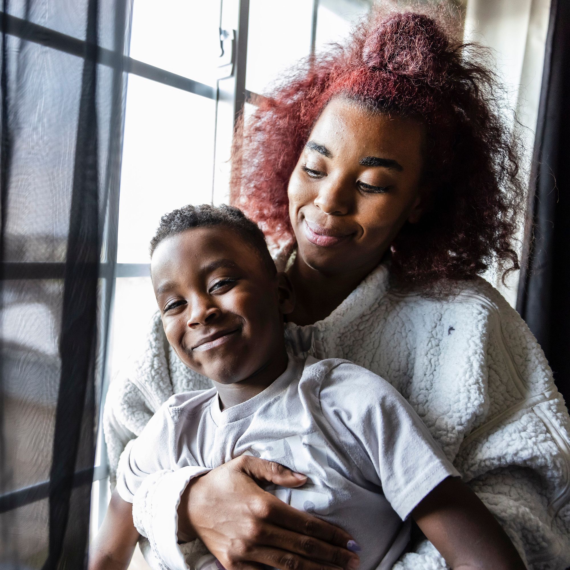 A portrait shows a Black woman, with red hair and a white sweater, smiling as she holds onto her young son. 
