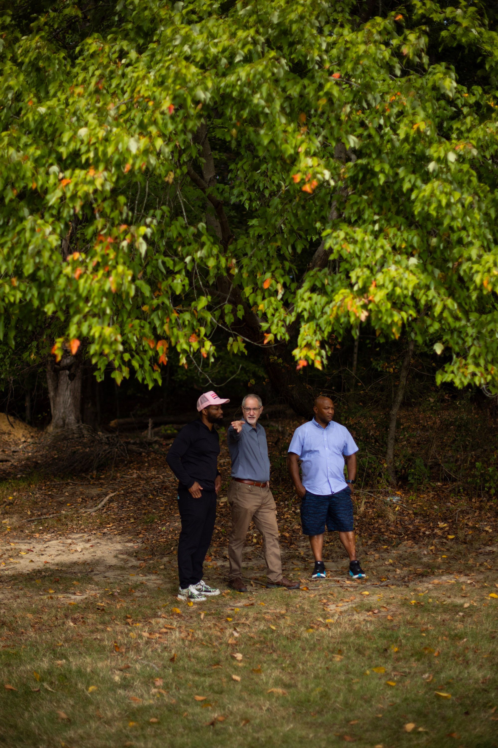 A photo of a White man pointing next to two Black men. The men are standing underneath a tree.