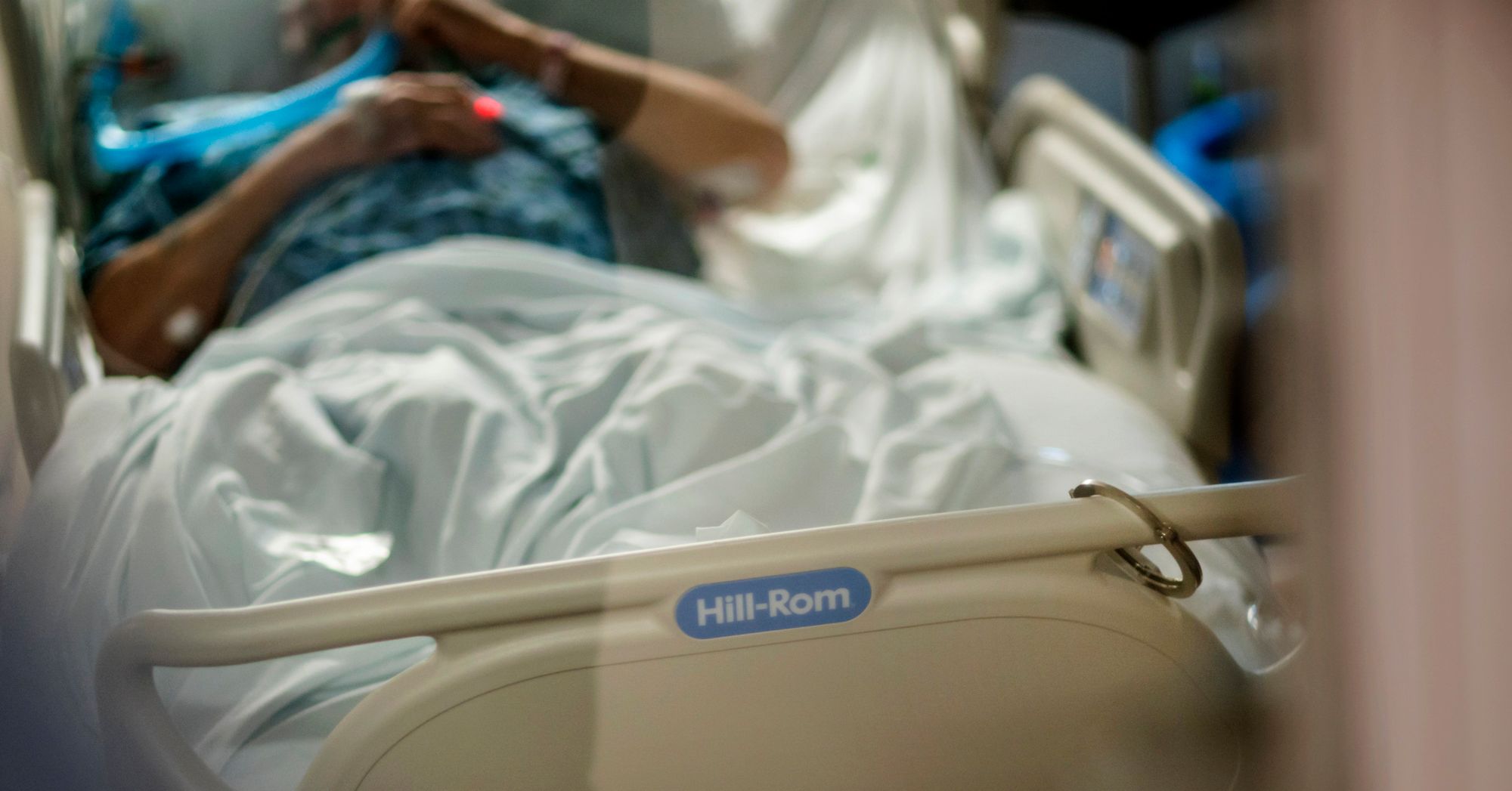 A person wearing a blue medical gown is holding a breathing tube while lying on a hospital bed. A handcuff is locked onto a bar of the bed.