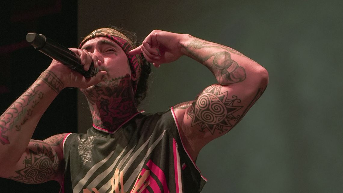 A photo shows a light-skinned man holding a microphone in his right hand while pointing to his head with his left hand. He is tattooed, and wearing a sleeveless jersey and a bandana. 