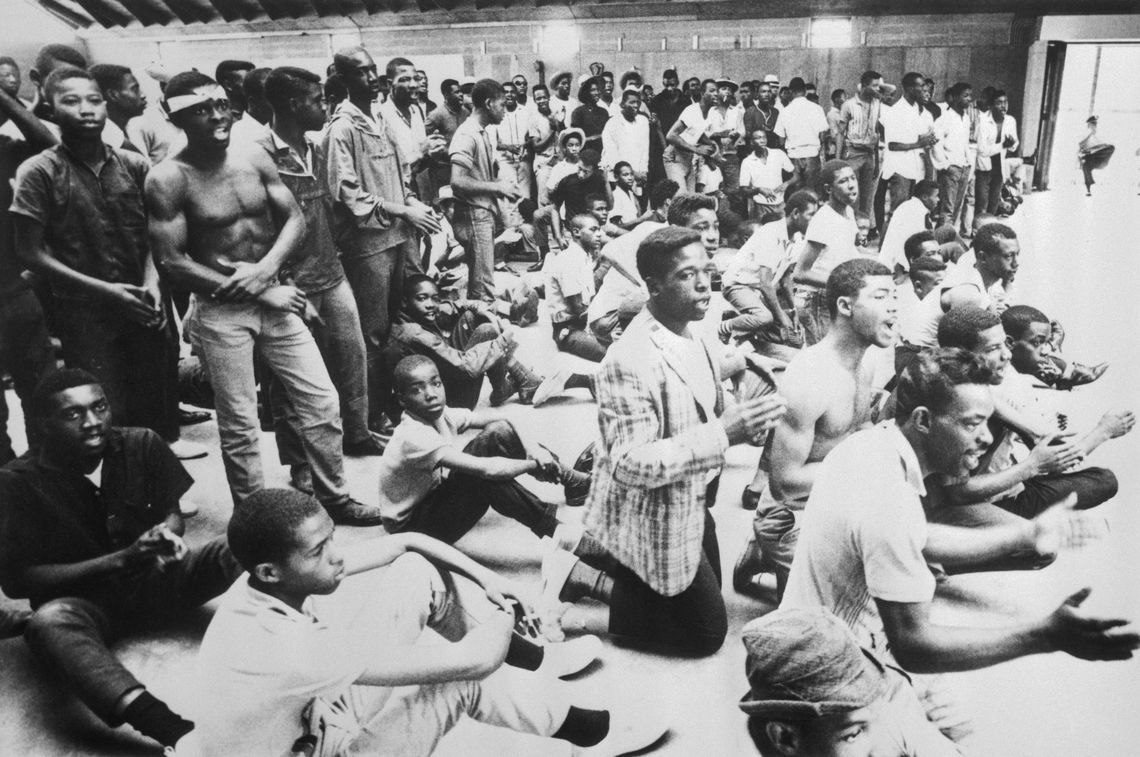 A black-and-white photograph shows a room full of Black men, some standing, some sitting or kneeling, singing and chanting. 