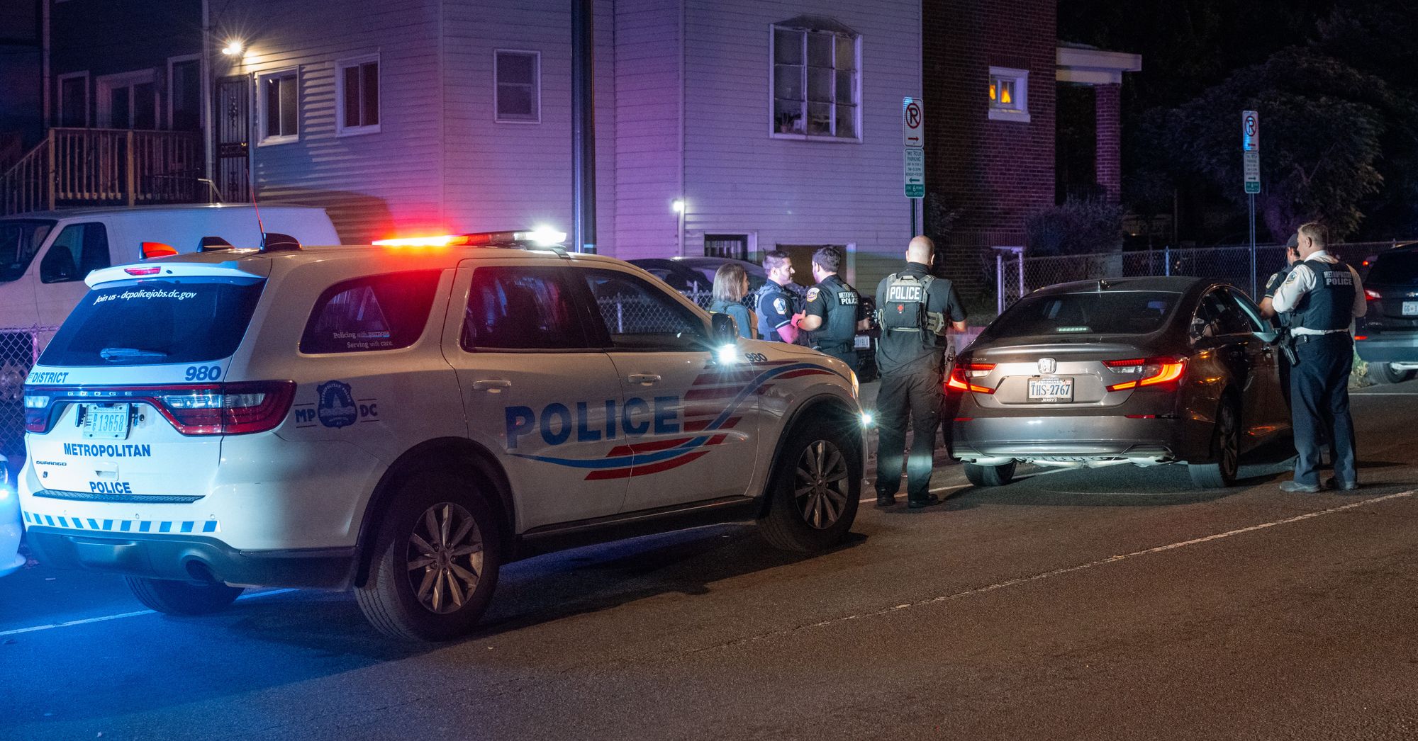A white police SUV is parked behind a gray car during a traffic stop. Four police officers stand the left side of the car while two officers stand on the right. 