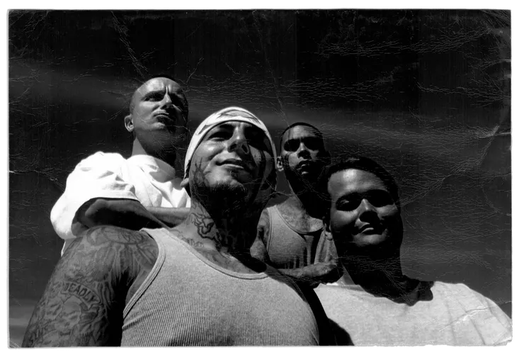 In a black and white photograph, Grace Pinson, a trans Latina with short dark hair, stands outside in the sun with three men. The person to her left wears a tank top and has tattoos on his face, neck and arms. Behind her stand two men. The one on the right wears a white T-shirt and is bald. The one on the left has tattoos on his chest and arms and has close-cropped, dark hair. 