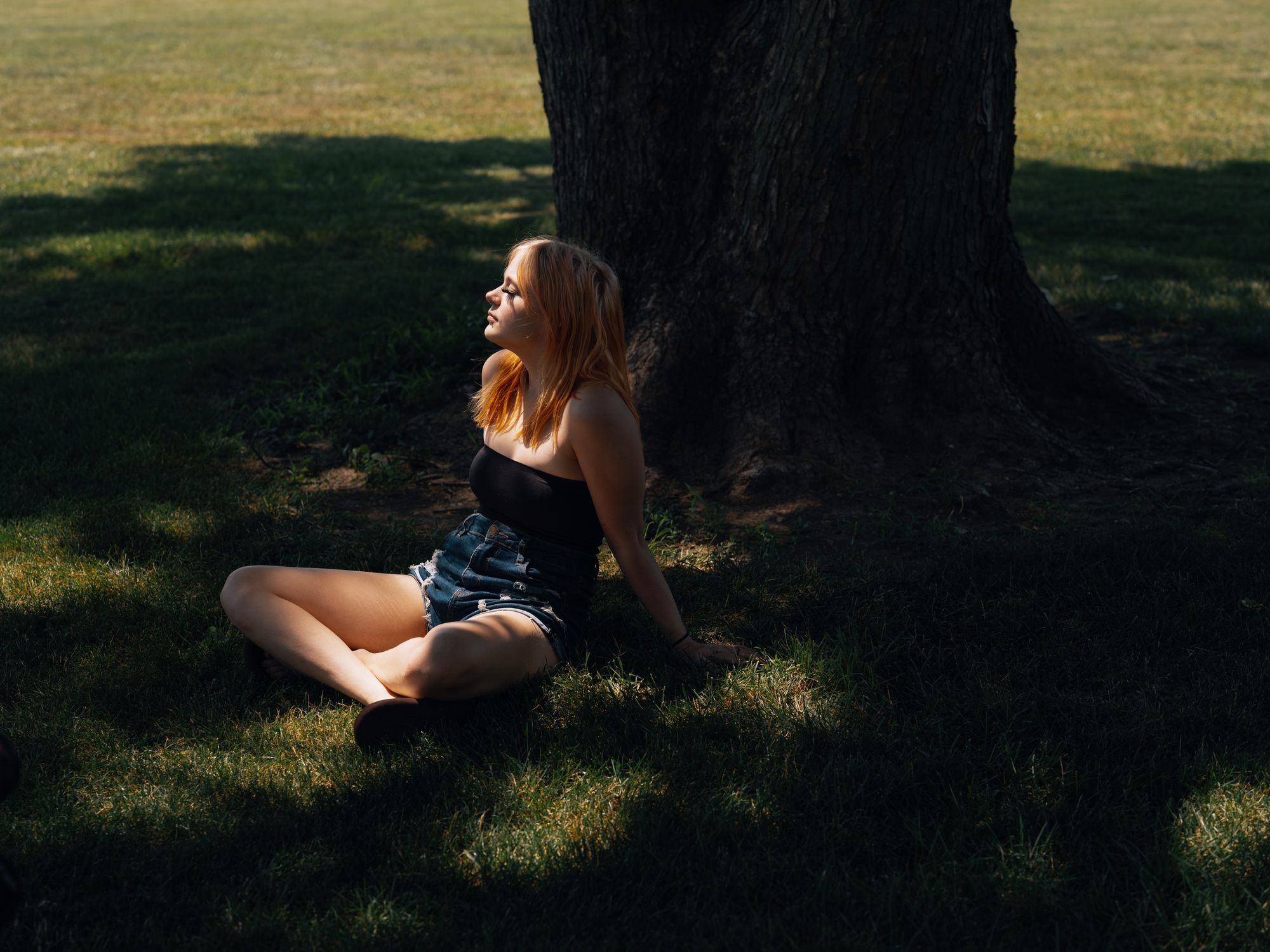 A White woman with reddish blonde hair, wearing a black tube top and jean shorts, sits under a tree. 