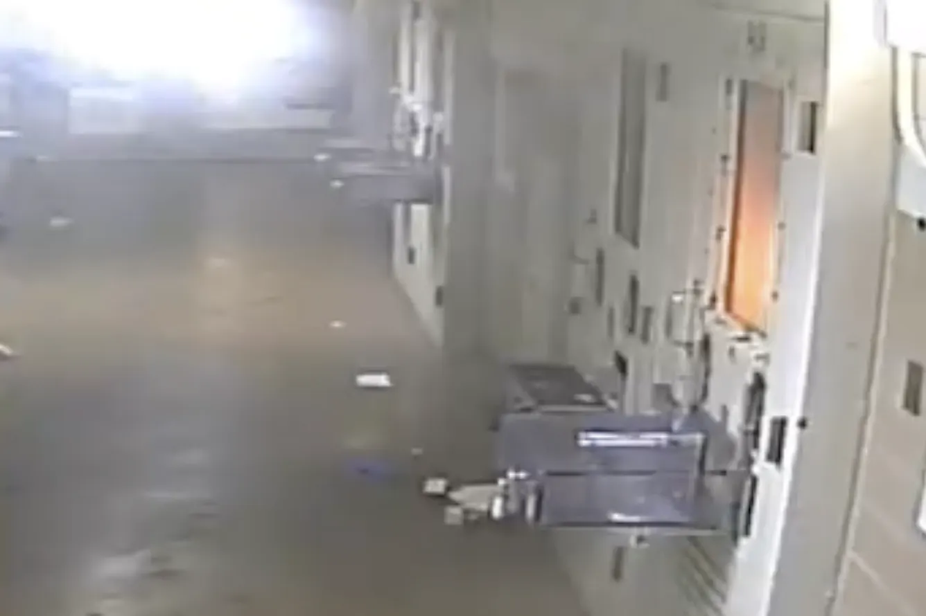 A video still shows a cell door in a prison hallway. A fire can be seen inside through the cell's window. 