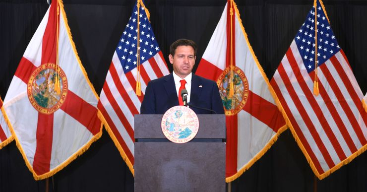 Florida Gov. Ron DeSantis, a White man in a navy suit and red tie, speaks into a microphone while standing at a podium with the seal of Florida. Flags of the United States and the state of Florida are in the background. 